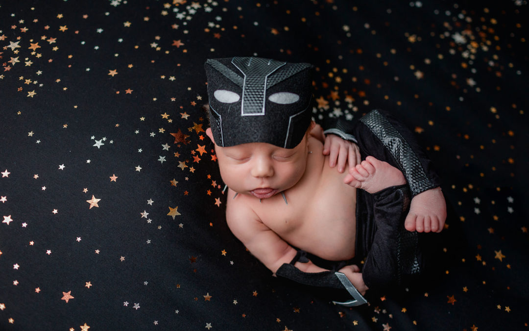 Black Panther Inspired Newborn Studio Pictures of baby boy laying on a black backdrop with golden stars wearing a black panther newborn outfit