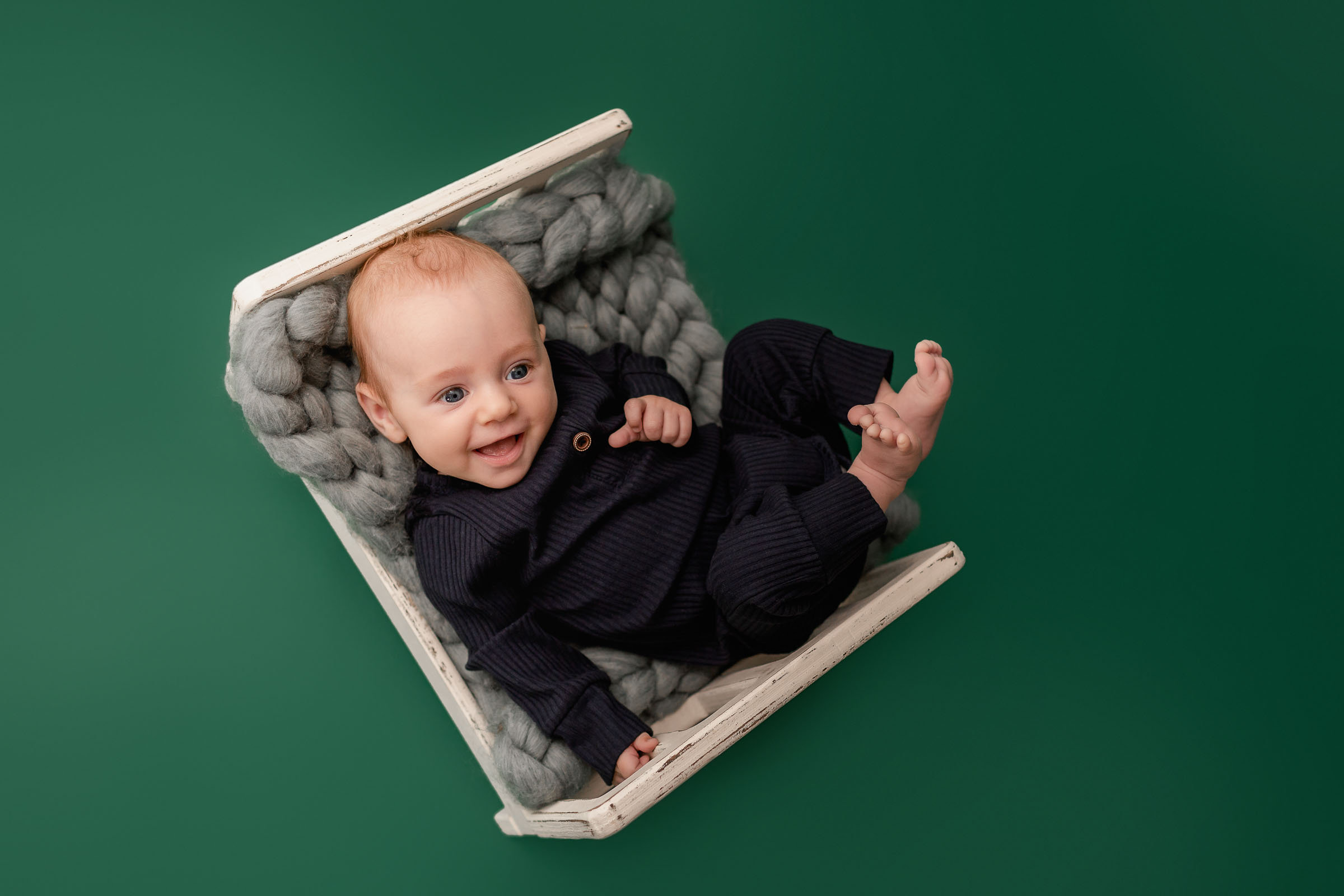 Happy 3 month old baby boy in blue outfit on bed on green backdrop