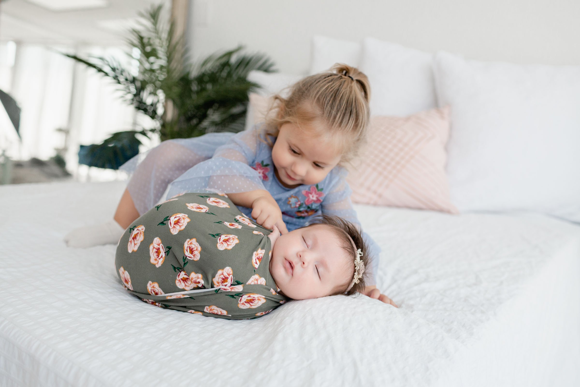 Picture of older sibling poking little sibling on the cheek during newborn pictures by Luxury Newborn Photographer Studio Freyja in San Diego. Big sister is slightly out of focus with newborn sister in focus wrapped up in a flower wrap.