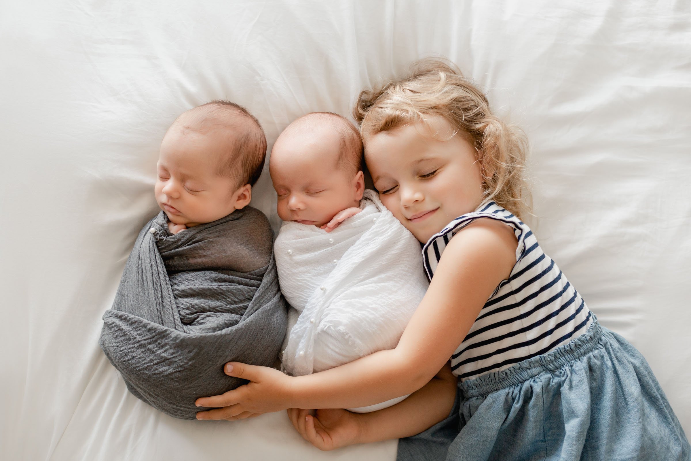 Older sister is posing with her twin newborn brothers during a newborn photoshoot in San Diego. They're all laying on a bed with older sibling hugging around the newborn twin brothers.