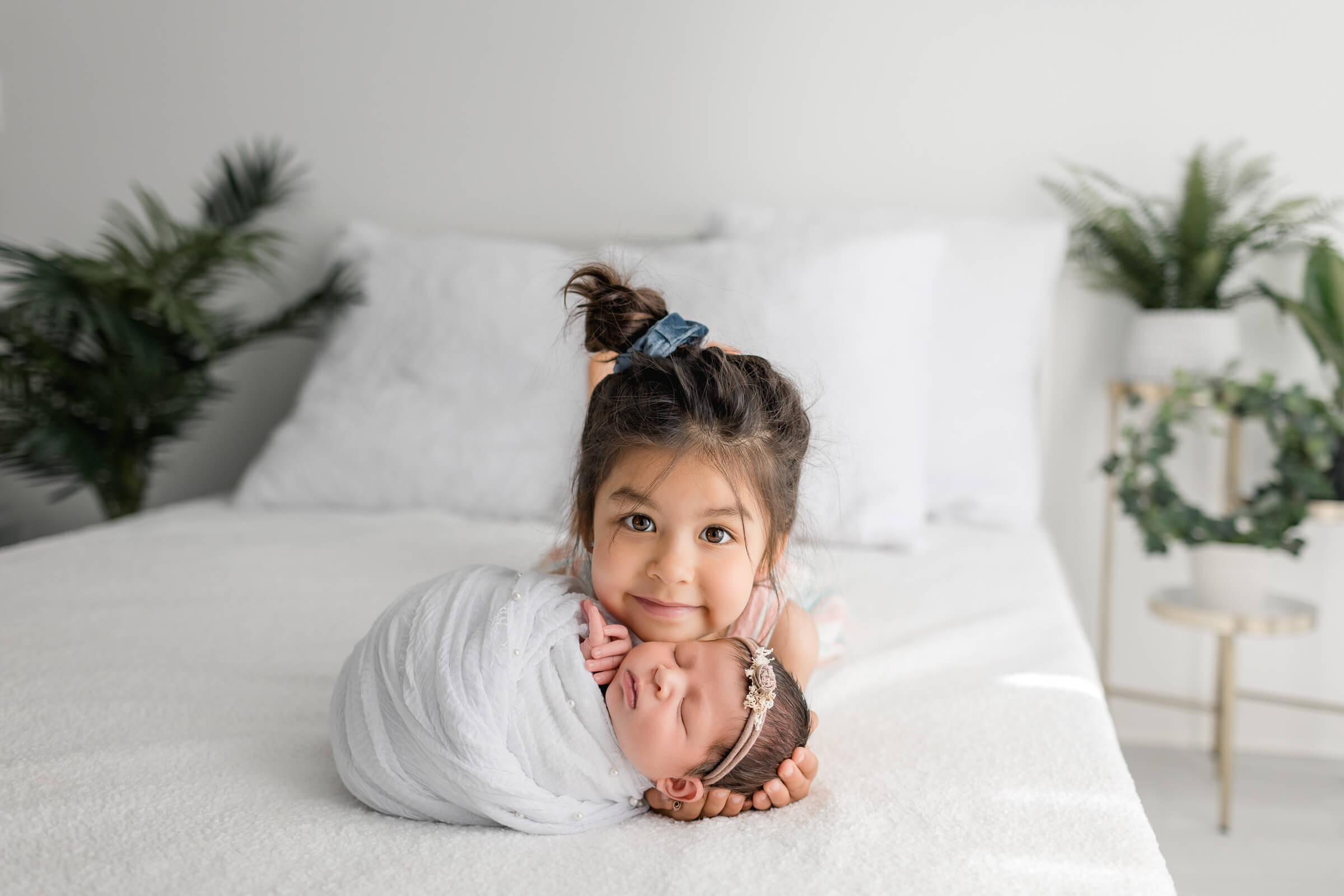Big Sister and Newborn Sister posing on a bed during newborn photos with newborn wrapped in white and big sister laying on stomach resting head on baby photographer by San Diego Newborn Photographer Studio Freyja
