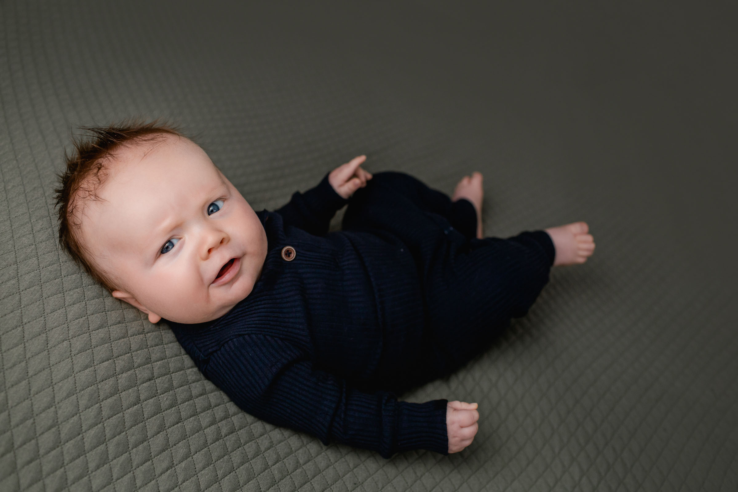 3 month old milestone photo with boy in dark blue outfit on green backdrop laying on his back looking at camera
