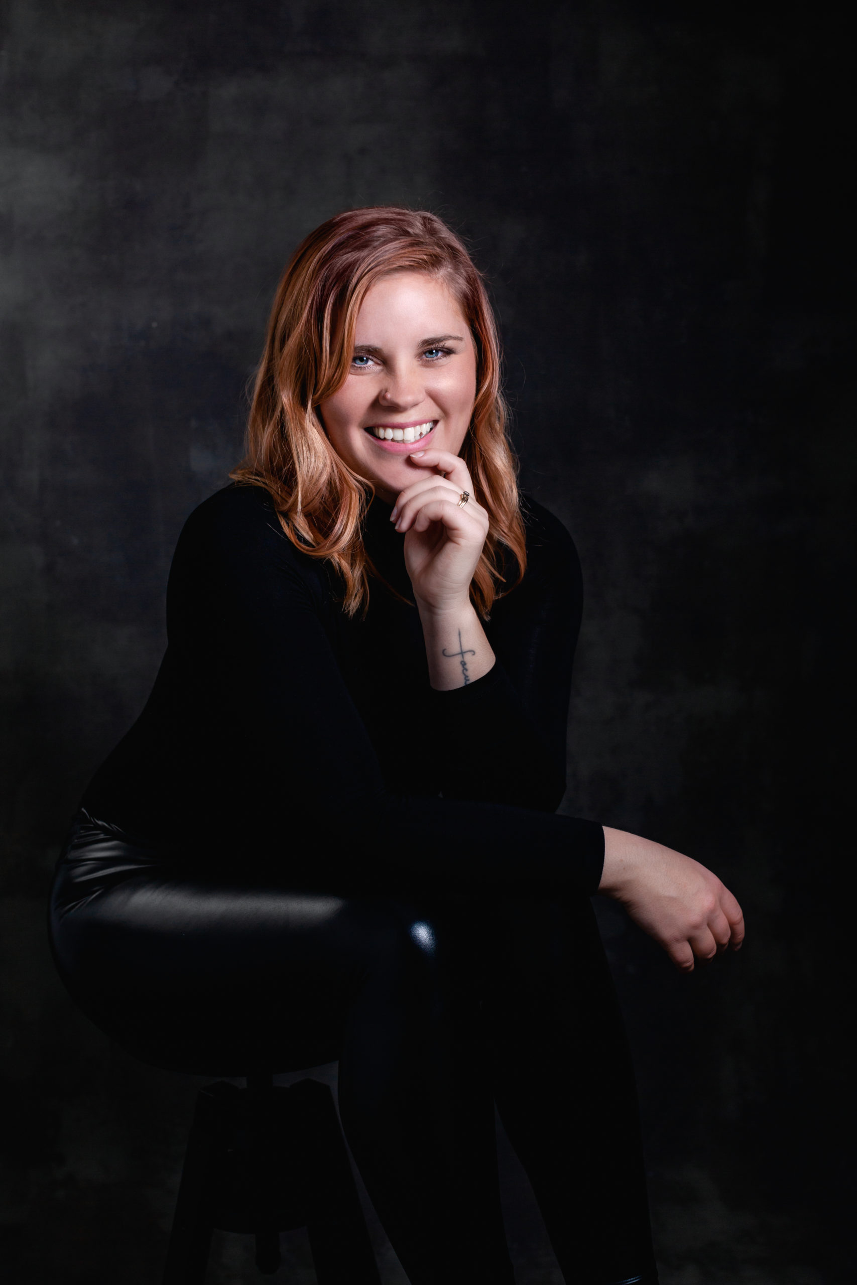 Headshot image of San Diego Photographer wearing black with black background. Photographer is sitting on a stool with knees bent and one hand on her chin smiling and looking at camera.