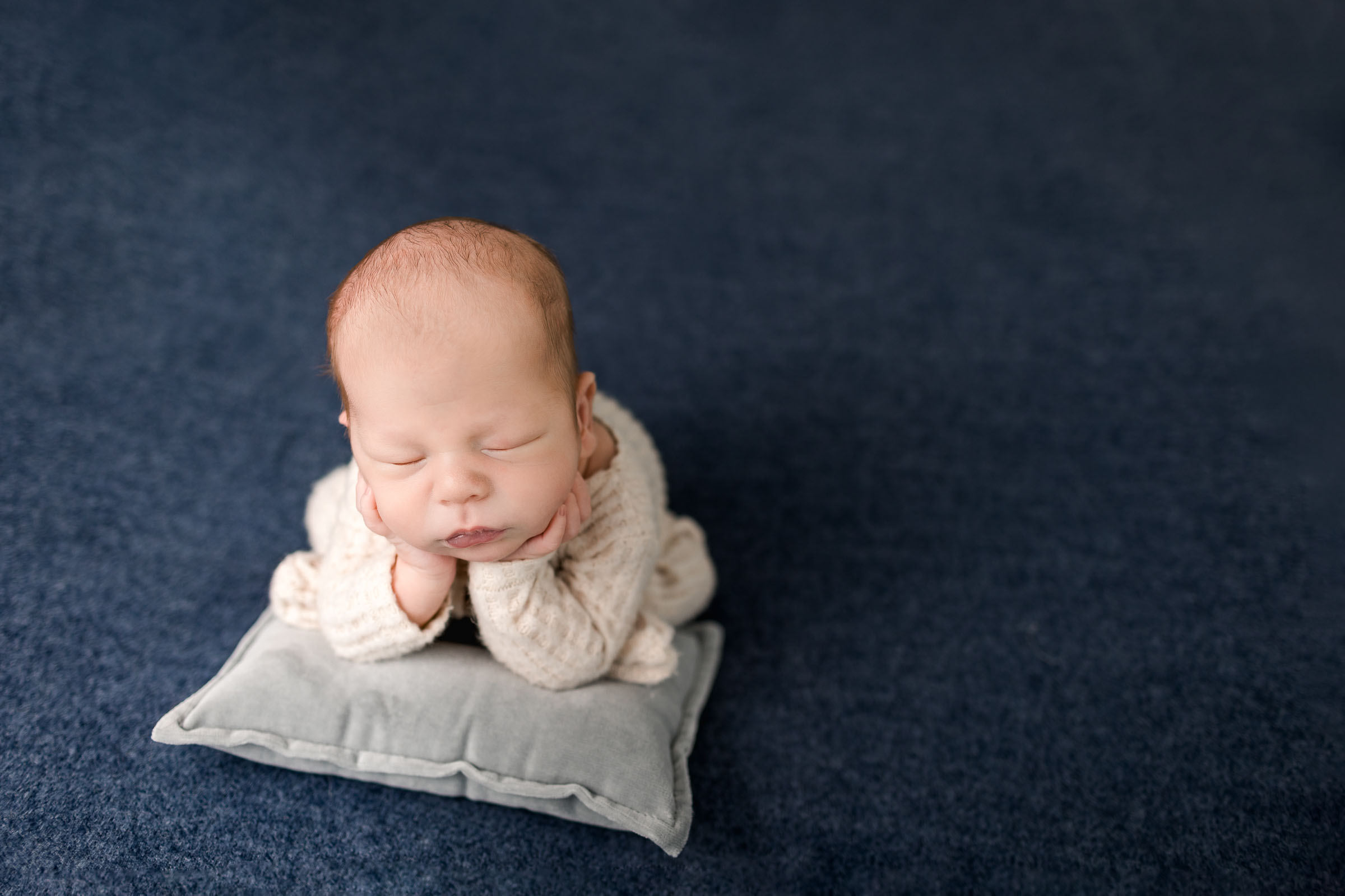 Sweet image of newborn boy in beige outfit doing froggy pose on a gray pillow with blue backdrop