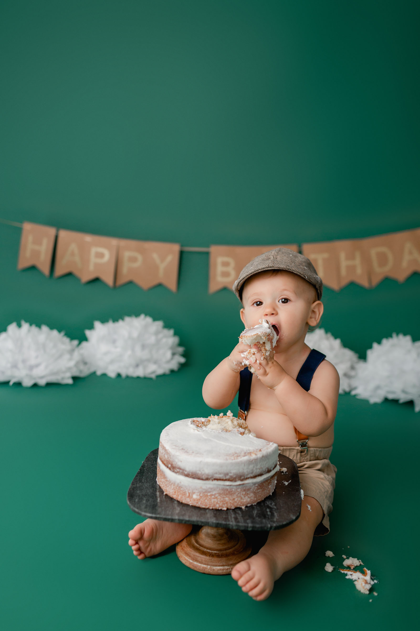 Boy in newspaper hat sitting on green backdrop eating his cake for his cake smash pictures