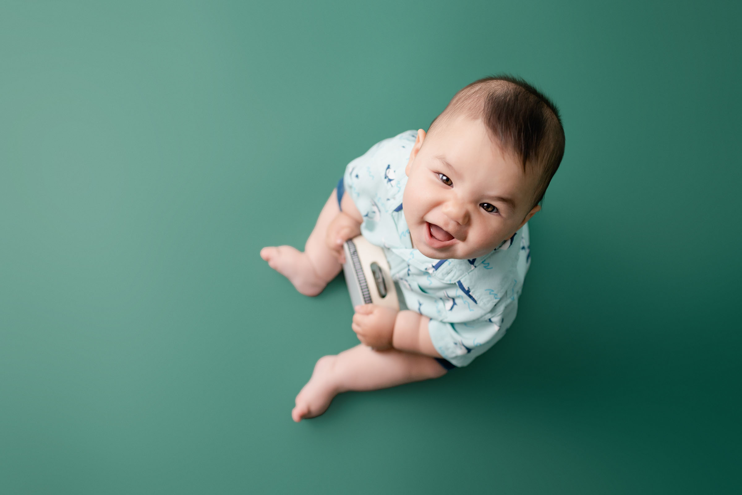 Super smiling baby boy sitting on green backdrop in blue shirt looking up at San Diego Baby Photographer