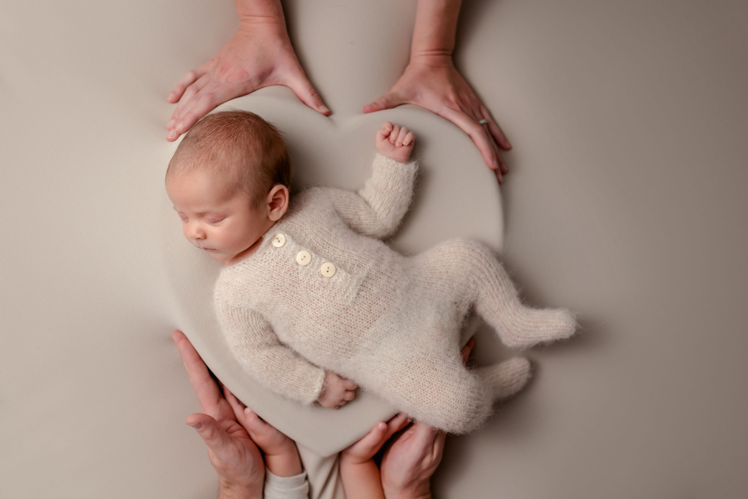 Neutral colored newborn photo with baby laying in a heart bowl surrounded by parents hands
