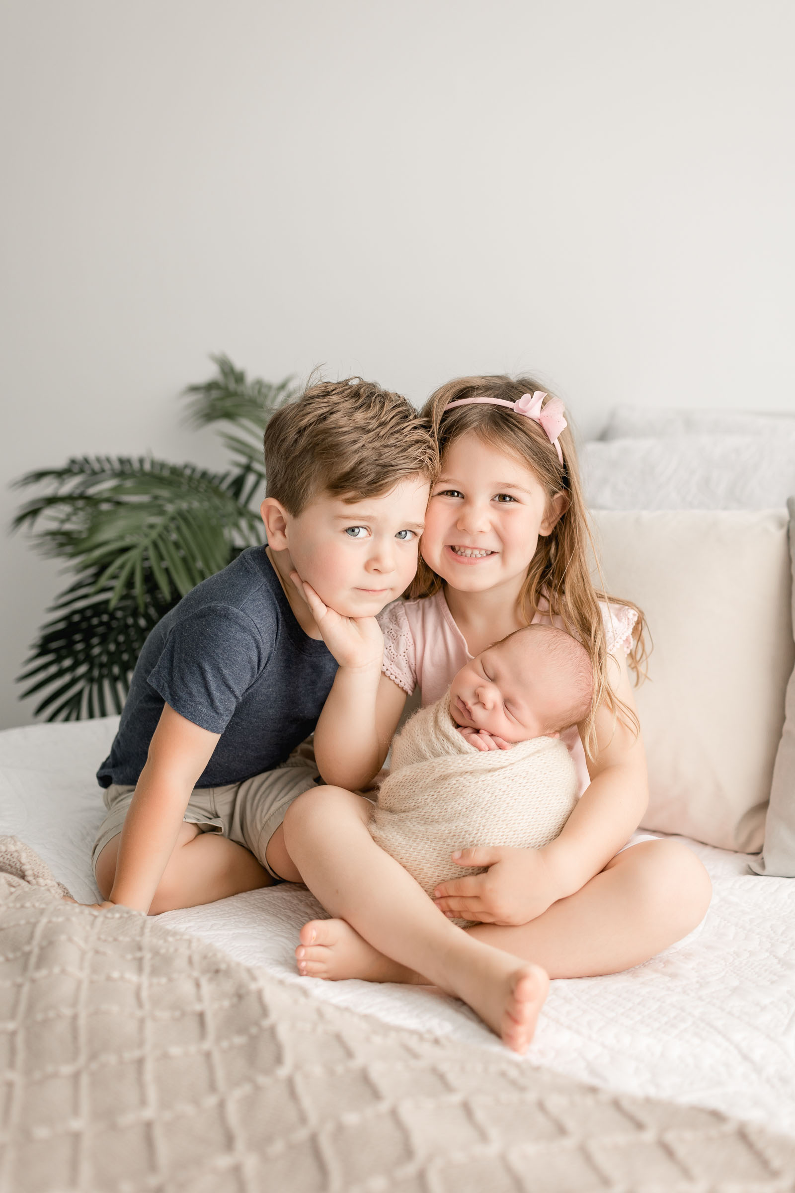Proud big sister holding newborn baby brother in lap with middle brother sitting next to her on bed in San Diego Photography Studio