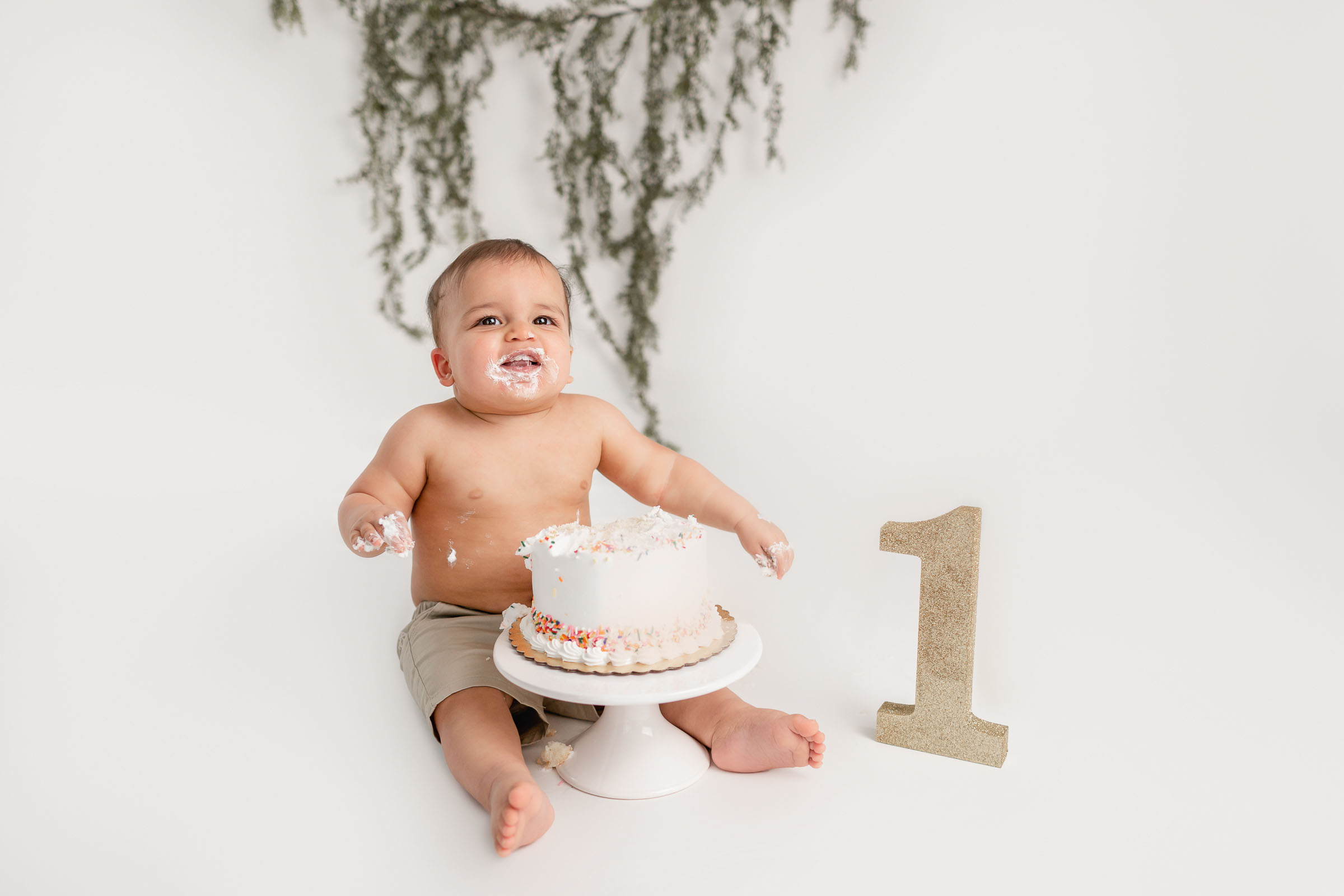 Boy in just shorts sitting behind his cake with cake on his face smiling for his cake smash pictures