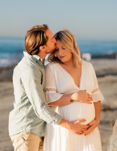 Dad giving pregnant mom a kiss on the head while holding belly during maternity photos in La Jolla