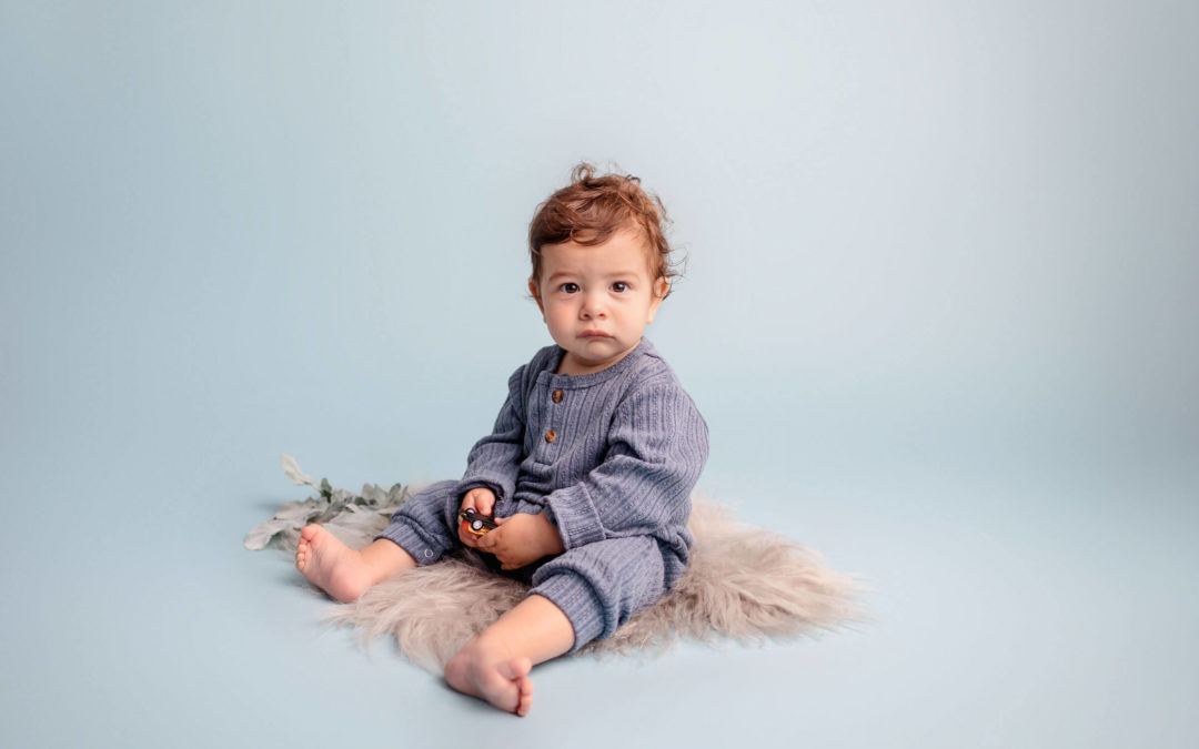 One Year Old Boy in Blue outfit sitting on a gray fur rug against a sky blue backdrop posing for his San Diego Photography Studio One Year Portraits