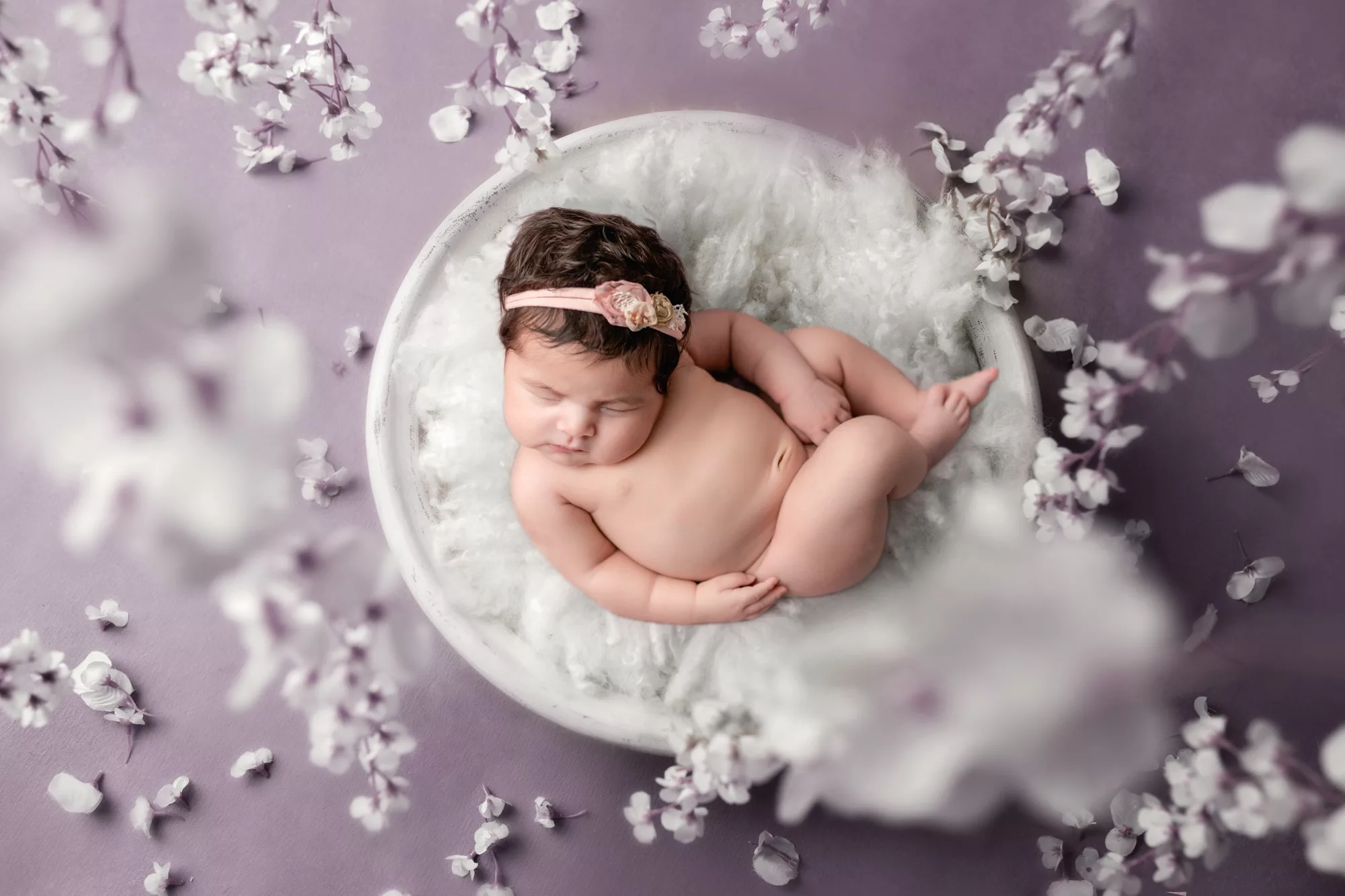 Picture of a newborn girl laying curled up in a white bowl with white fluff surrounded by white floral coming down from the ceiling