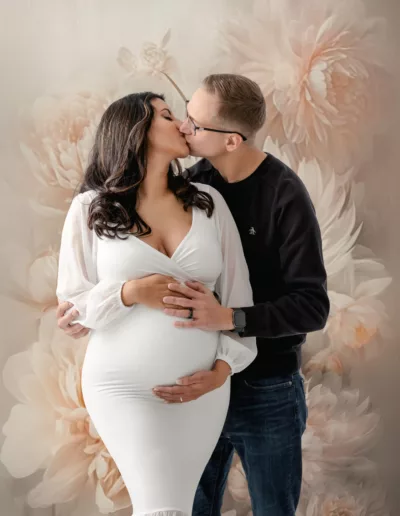 Expectant mom and dad standing in front of a floral backdrop cradling the stomach. Mom wearing a white maternity dress and dad wearing black