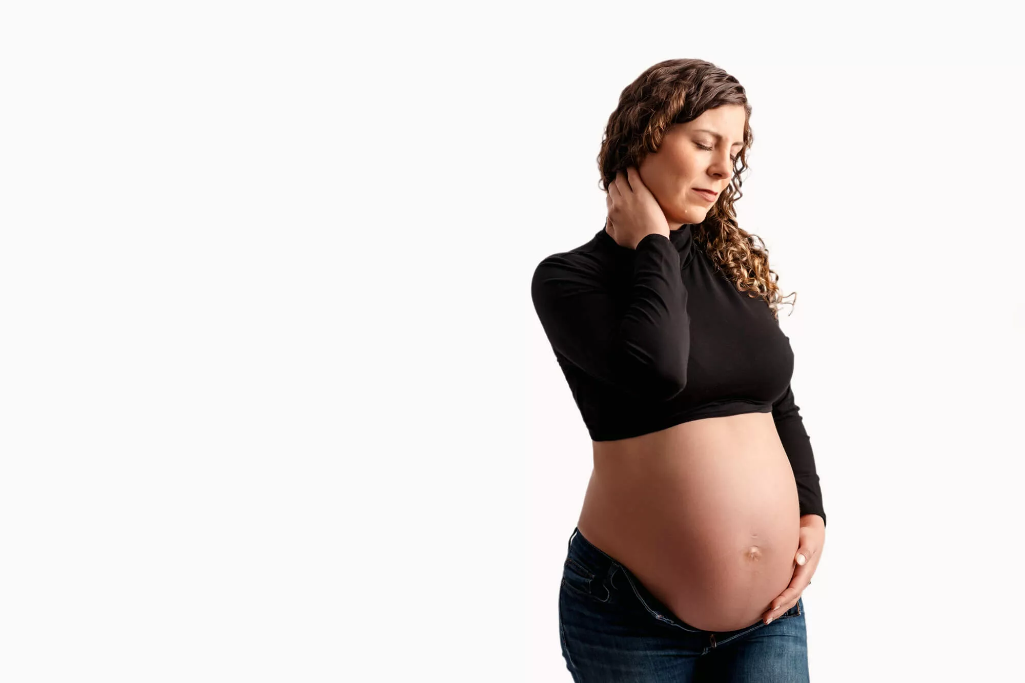 Elegant pregnant woman posing gracefully against a pristine white backdrop wearing a black crop top and jeans