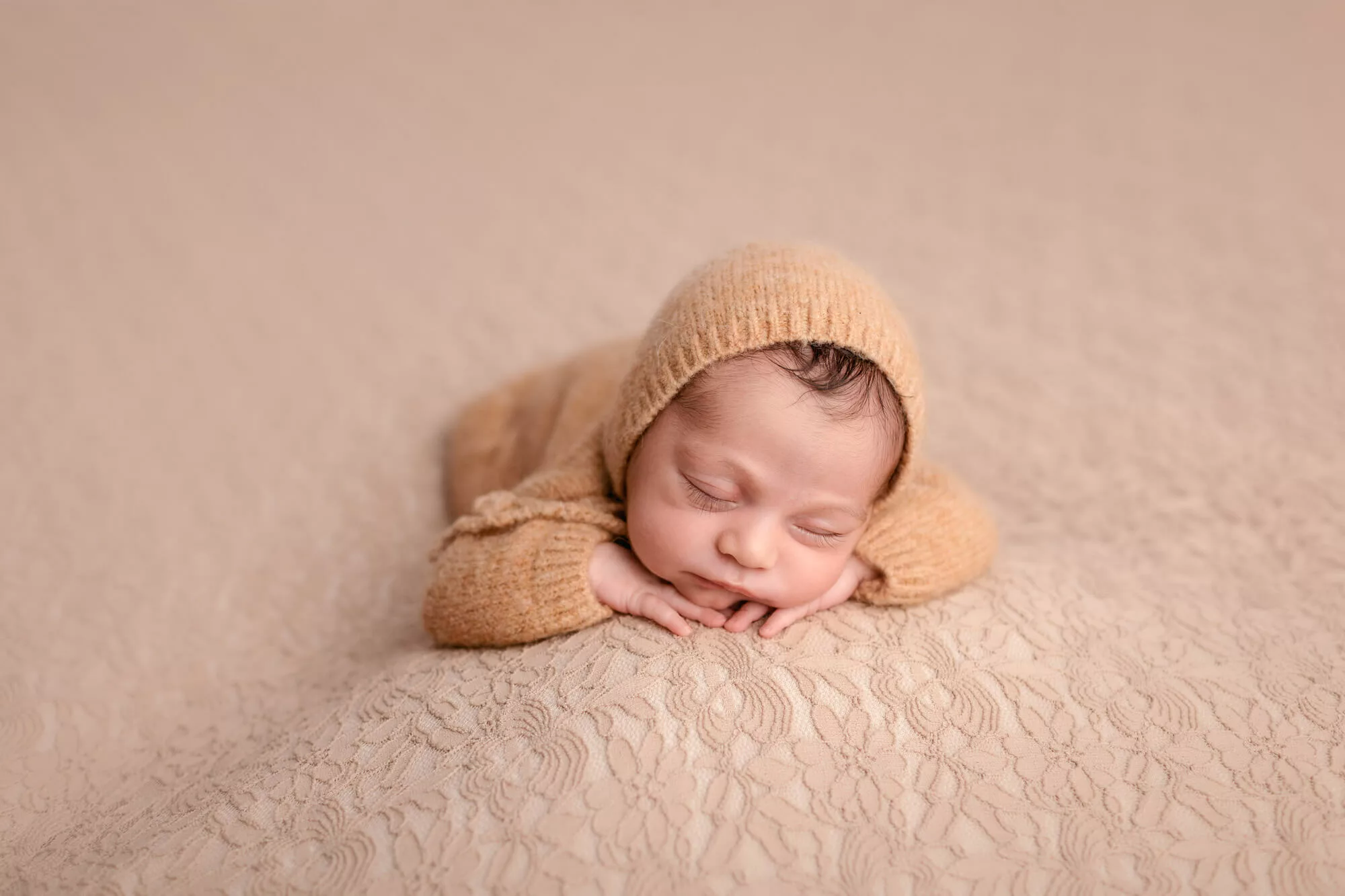 Newborn baby sleeping with heads on hands wearing a caramel colored oufit on a matching background