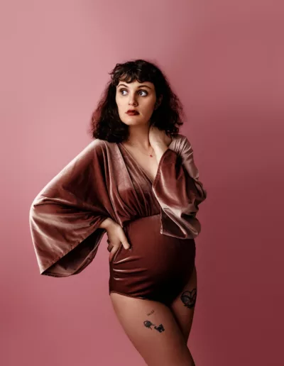 Pregnant woman wearing a pink velvet bodysuit against a pink backdrop showing off her baby bump