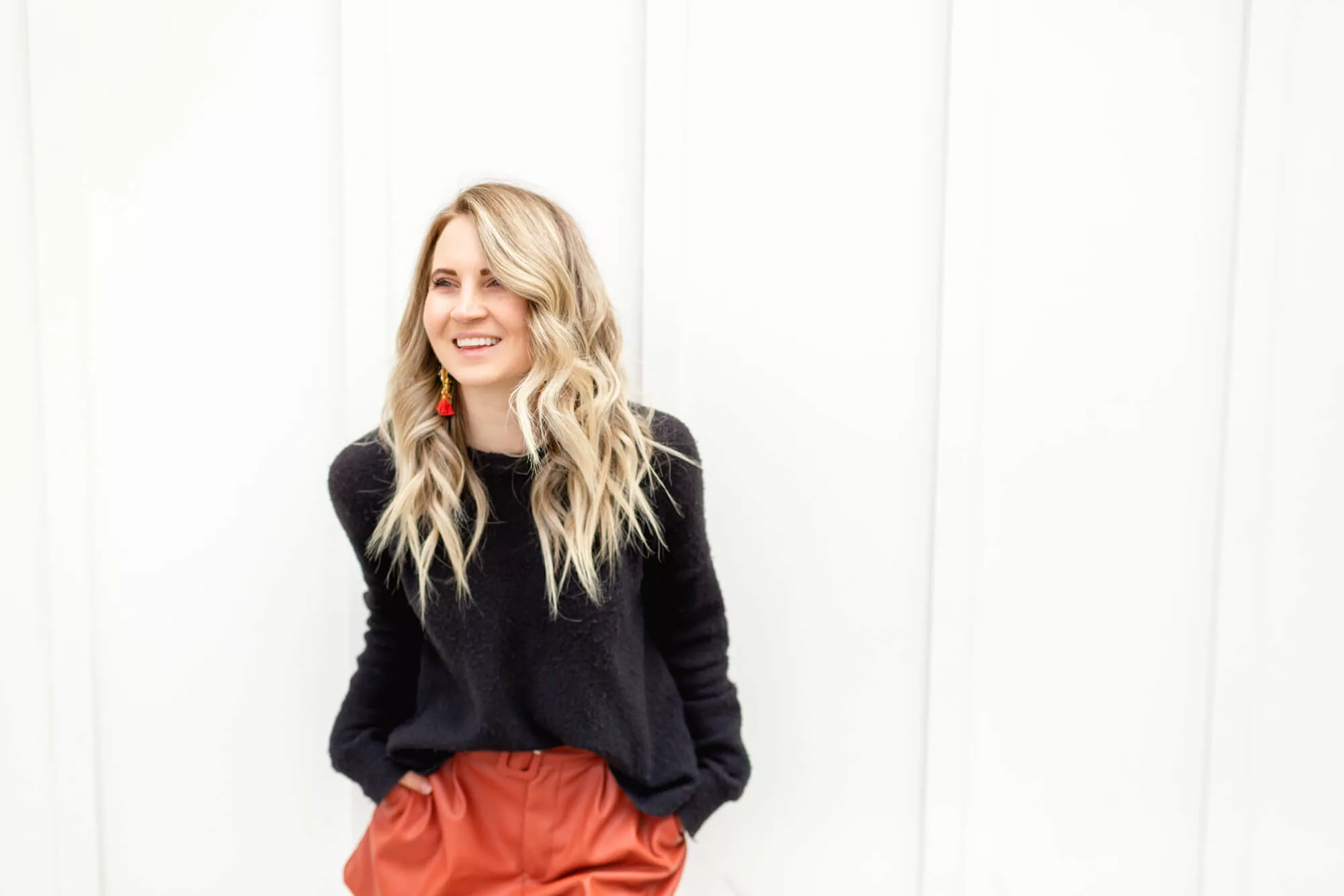 Elegant blonde woman in orange leather skirt and black sweater exudes sophistication and style during headshot session with San Diego Photographer. She's laughing while leaning up against a white wall.
