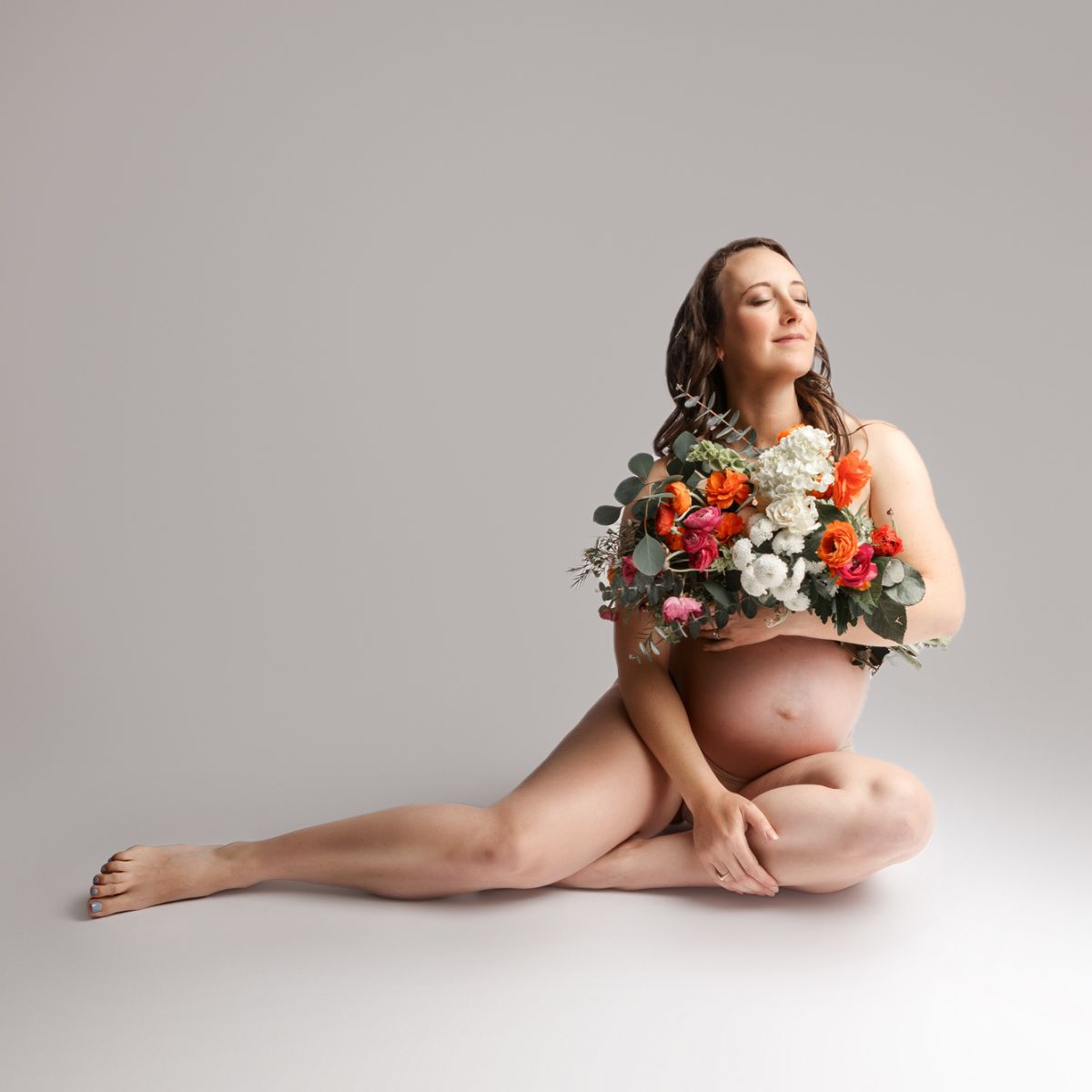 Fine art nude maternity portrait of mom against a white backdrop holding a bouquet of flowers capturing in San Diego Photography Studio