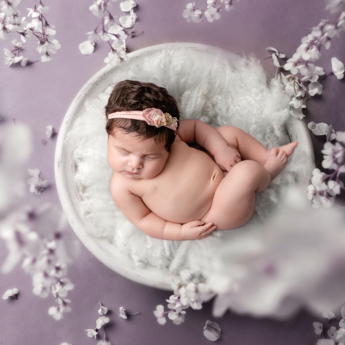Fine art studio newborn image of baby girl laying in a bowl with a purple backdrop and surrounded by white floral