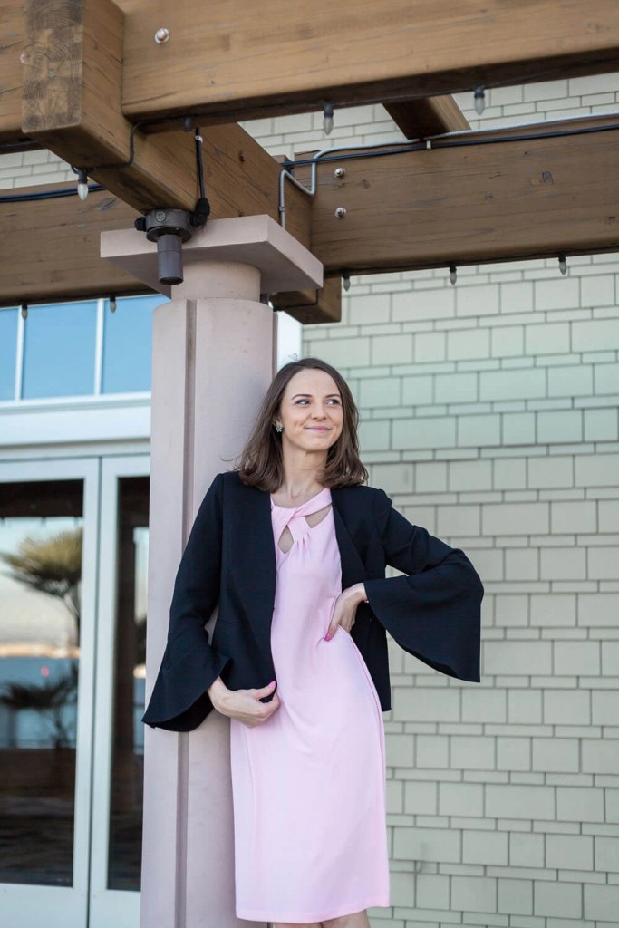 Real estate agent in a black blazer and pink dress leaning against a tan pillar, smiling