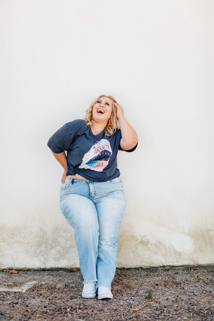 Life coach laughing joyfully against a white wall, hand in hair, casual denim outfit