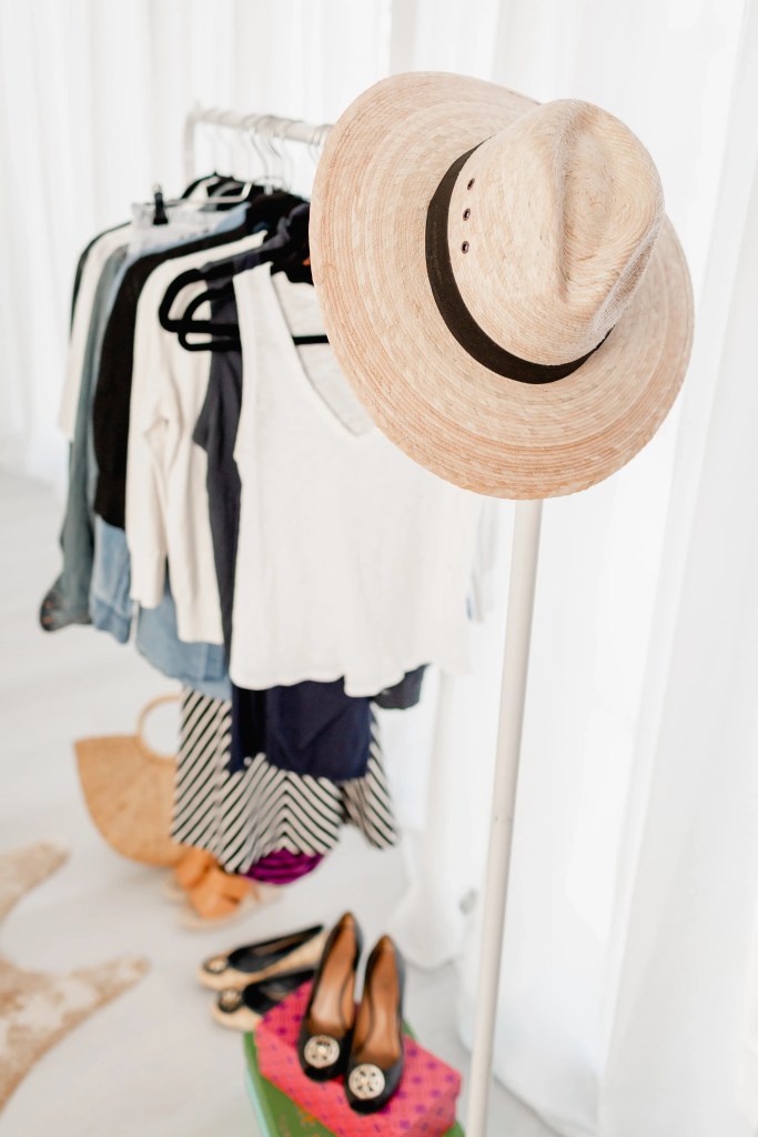Straw sun hat hanging on a clothing rack beside folded wardrobe pieces, styled studio flat lay