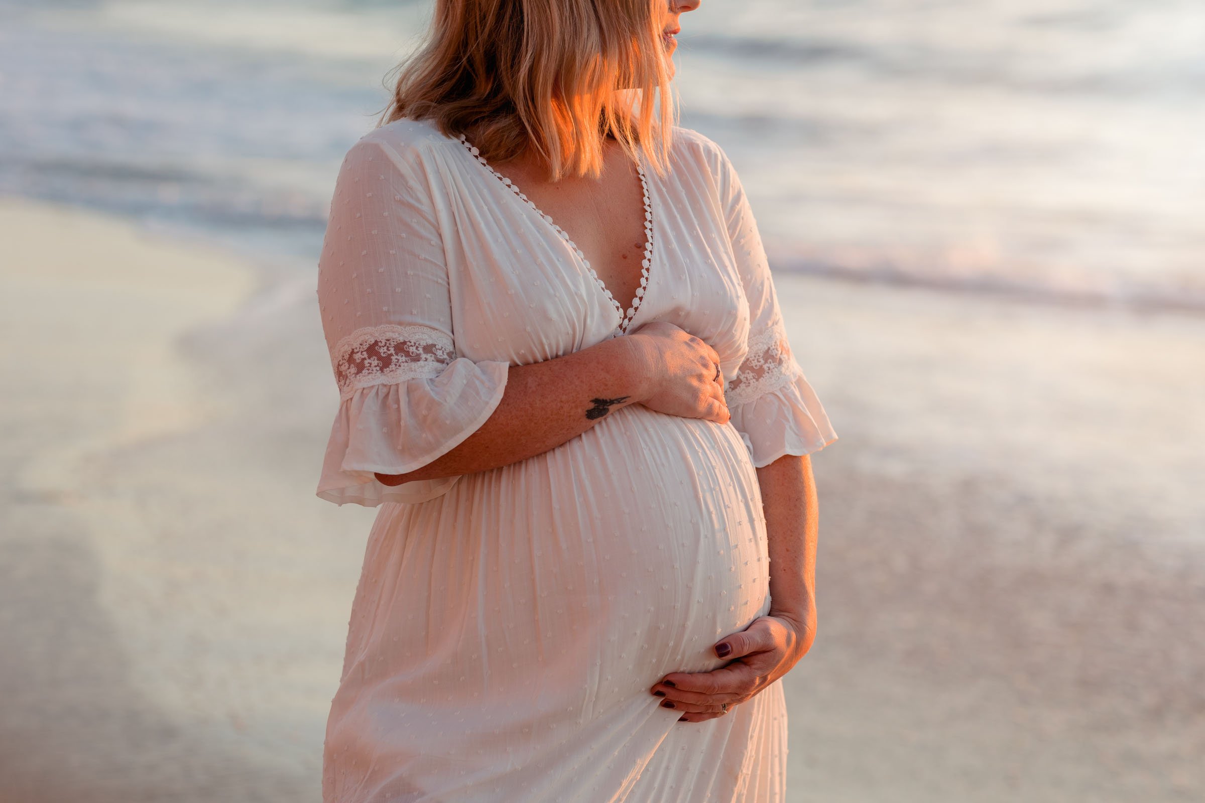 Pregnant woman in a white lace-sleeved dress cradling her bump on the beach at golden hour