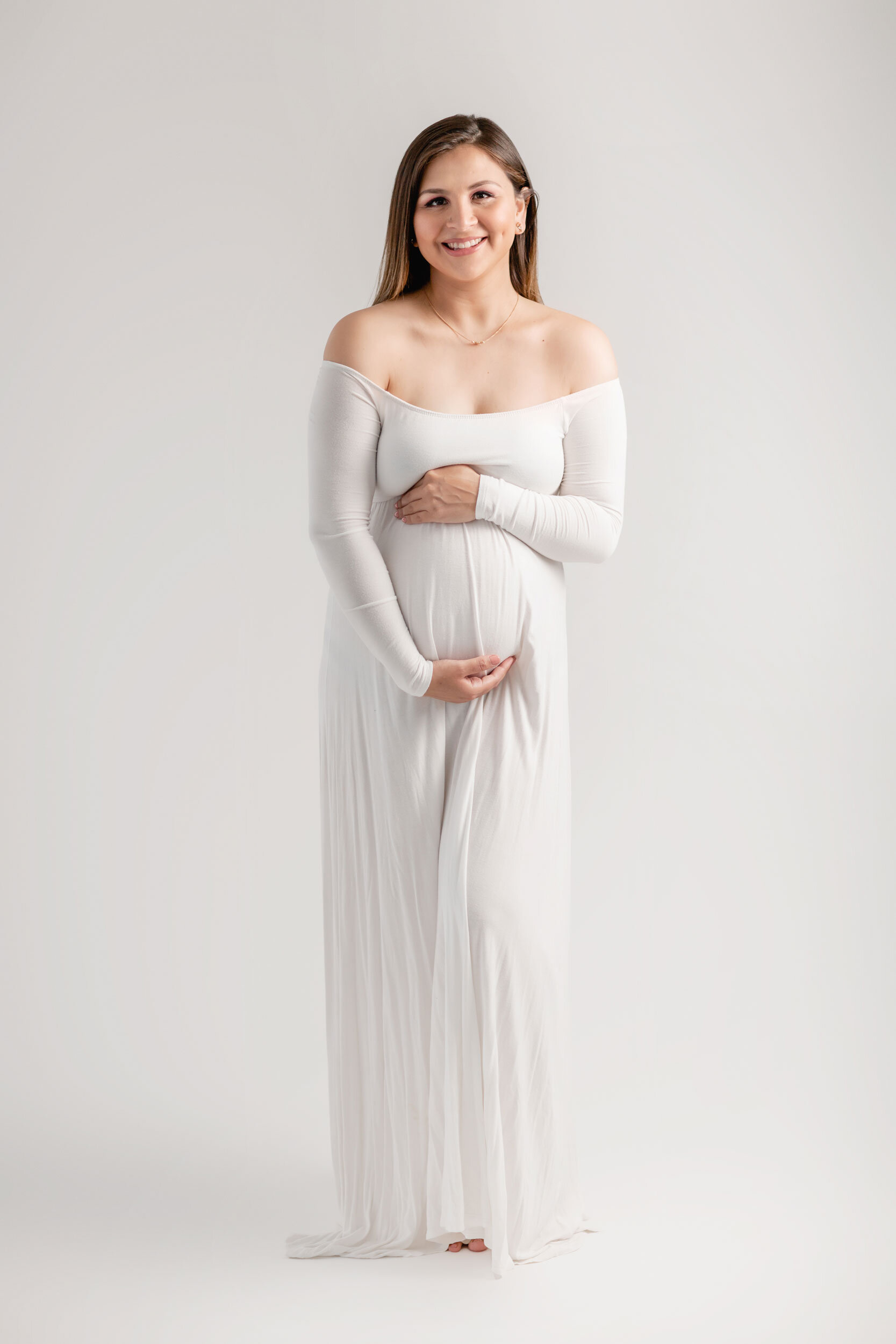 Smiling pregnant woman in a white off-the-shoulder gown on a light gray studio backdrop