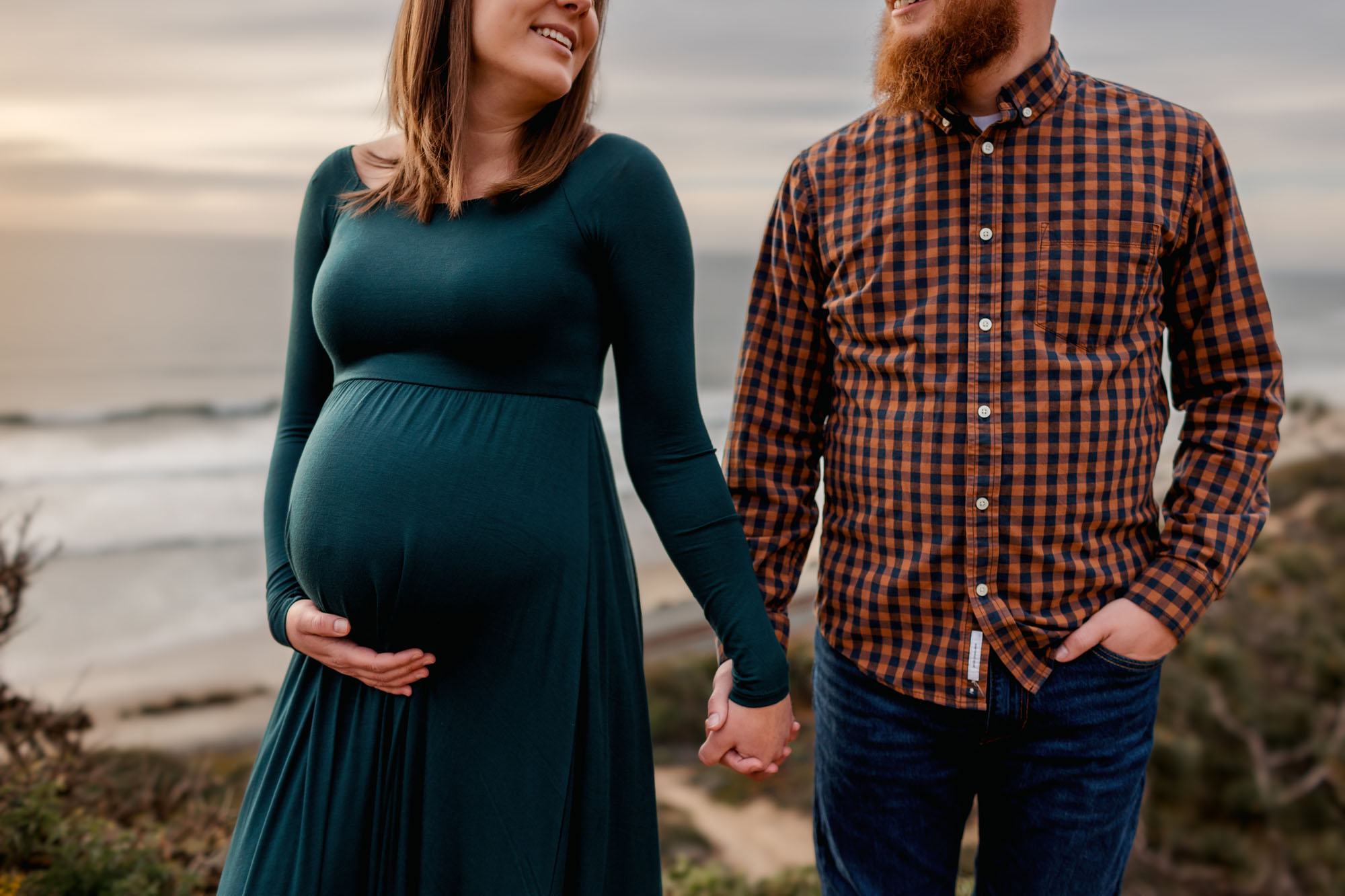 Expectant couple holding hands on a coastal bluff, she cradles her bump in a long green dress