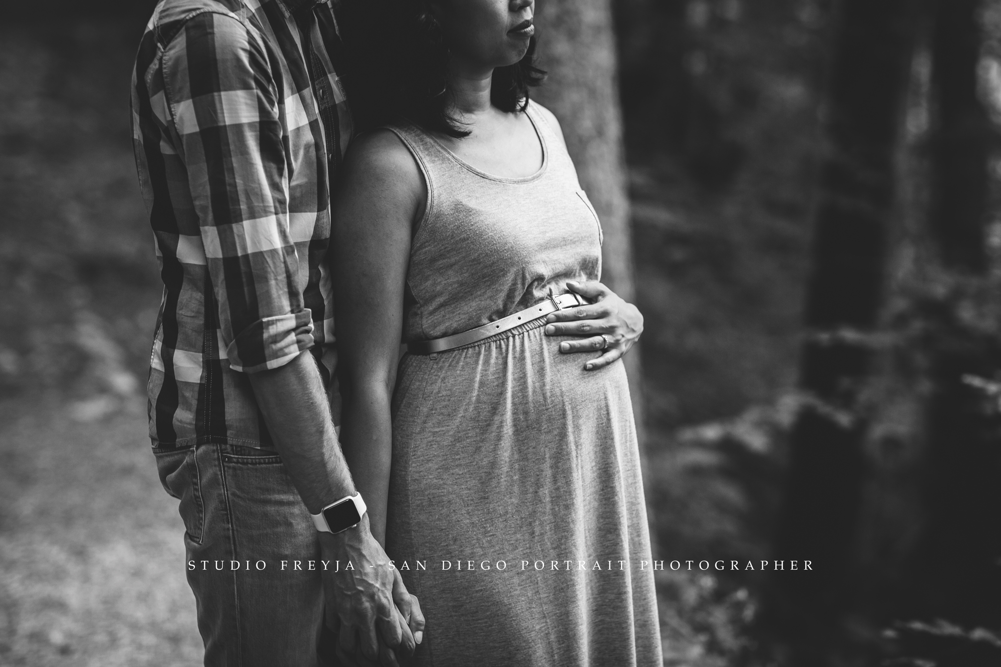 Black and white photo of an expectant couple side by side in a forest, her hand on her bump