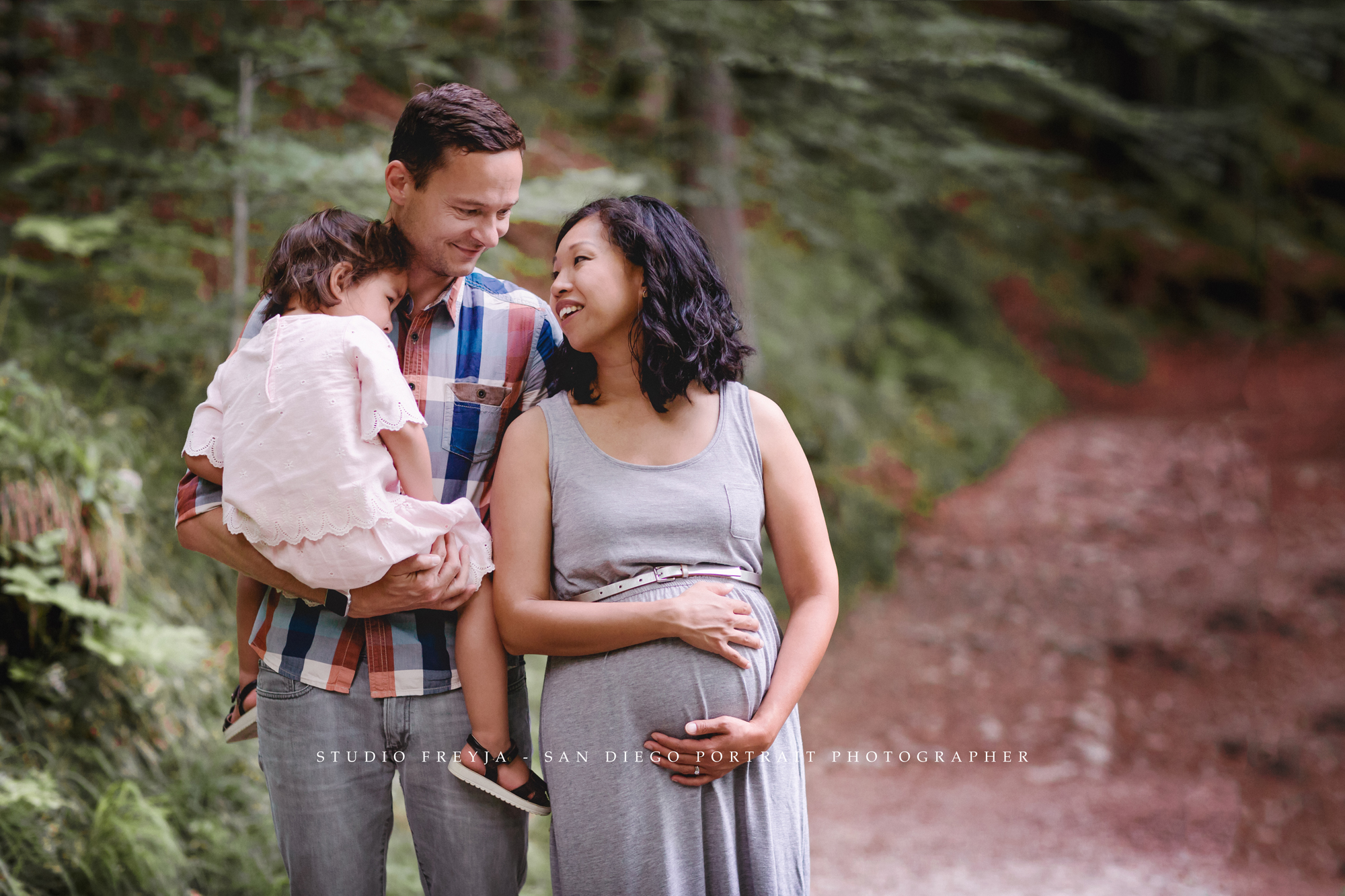 Expectant parents on a forest trail, dad holding their young daughter while mom cradles her bump