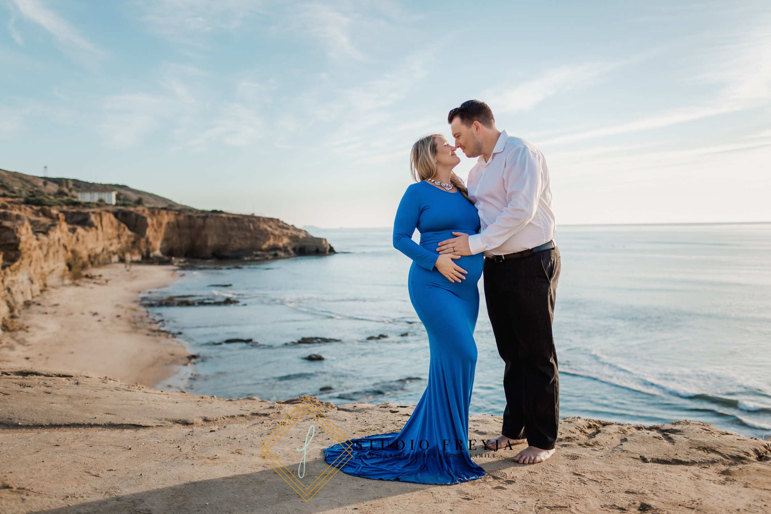 Expectant couple kissing on the cliffs at Sunset Cliffs, her royal blue gown trailing on the sand