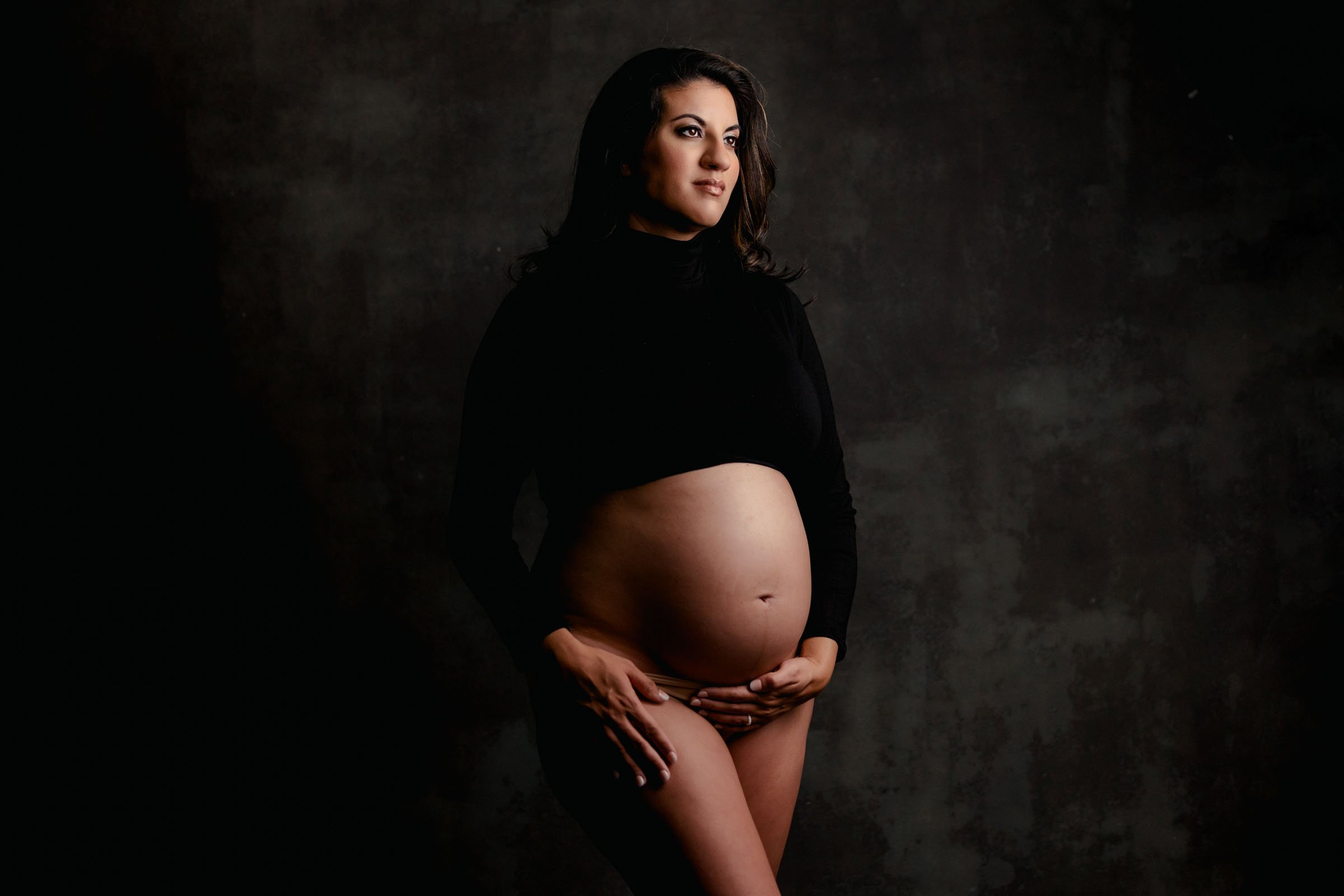 Pregnant woman in a black crop top showing her bump against a dark studio backdrop
