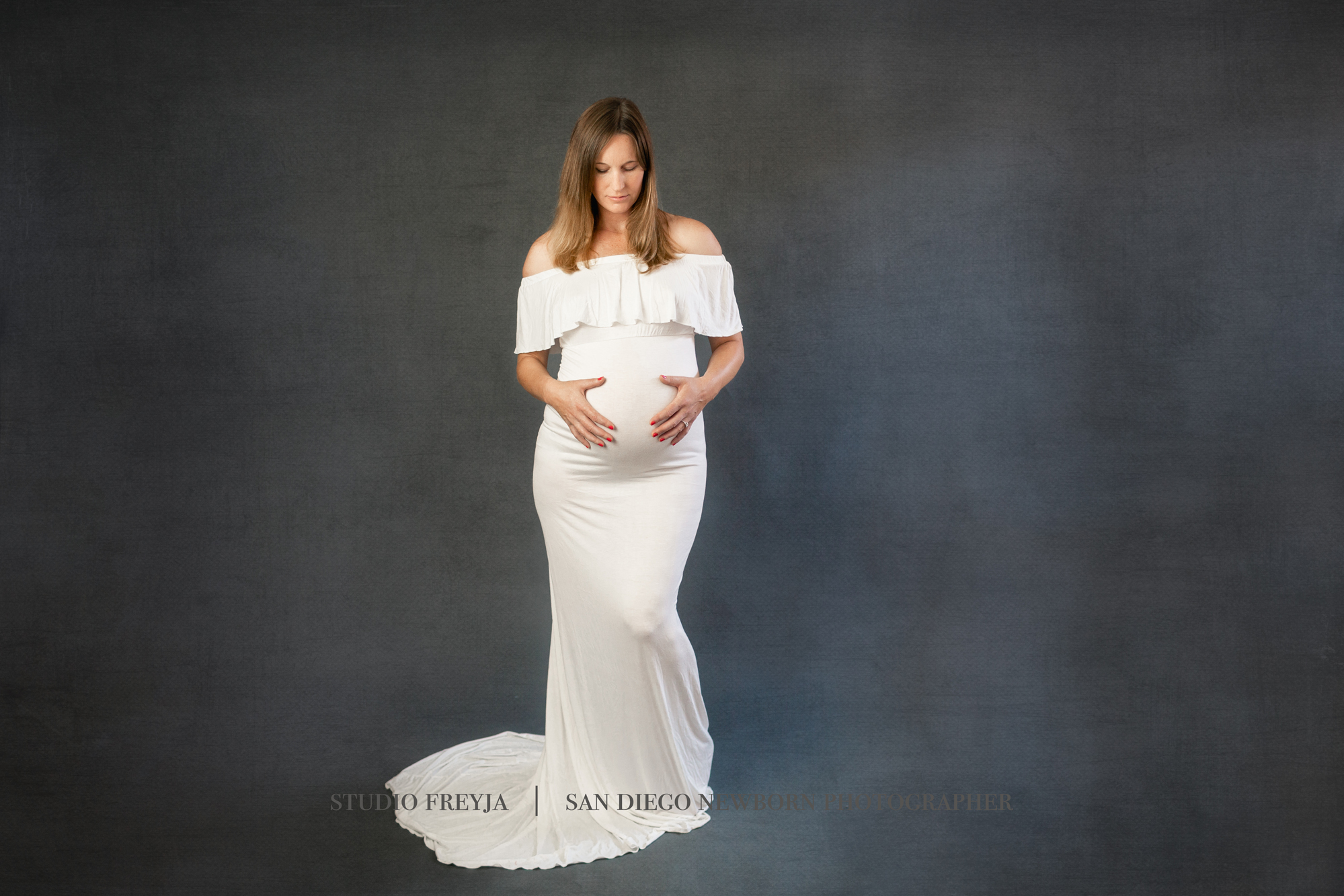 Pregnant woman in a white ruffled off-the-shoulder gown against a dark blue studio backdrop