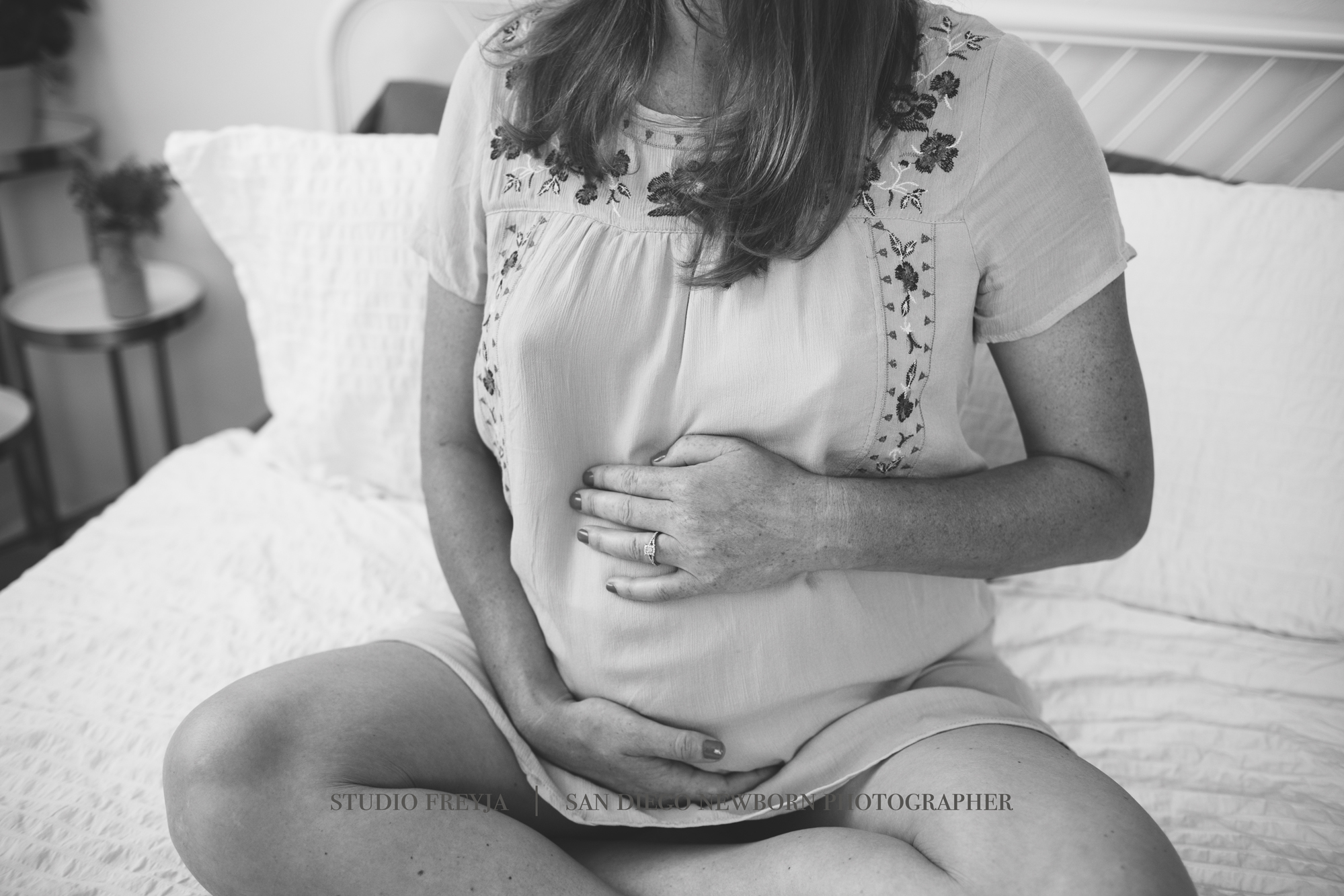 Black and white close-up of a pregnant woman sitting on a bed with her hands on her bump