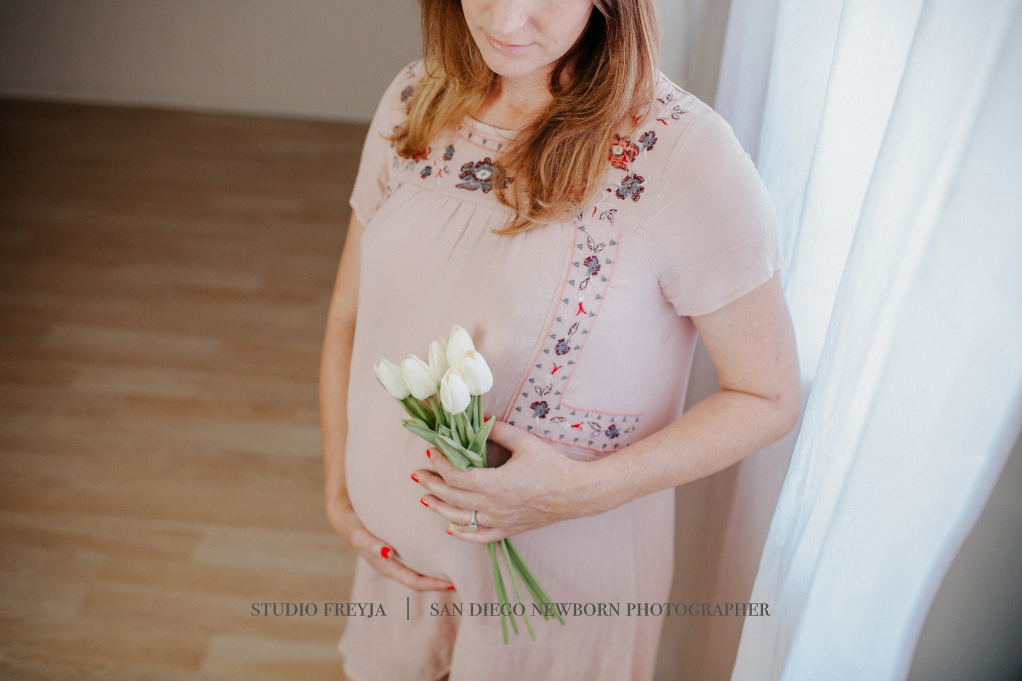 Pregnant woman in a blush embroidered dress holding white tulips by a window