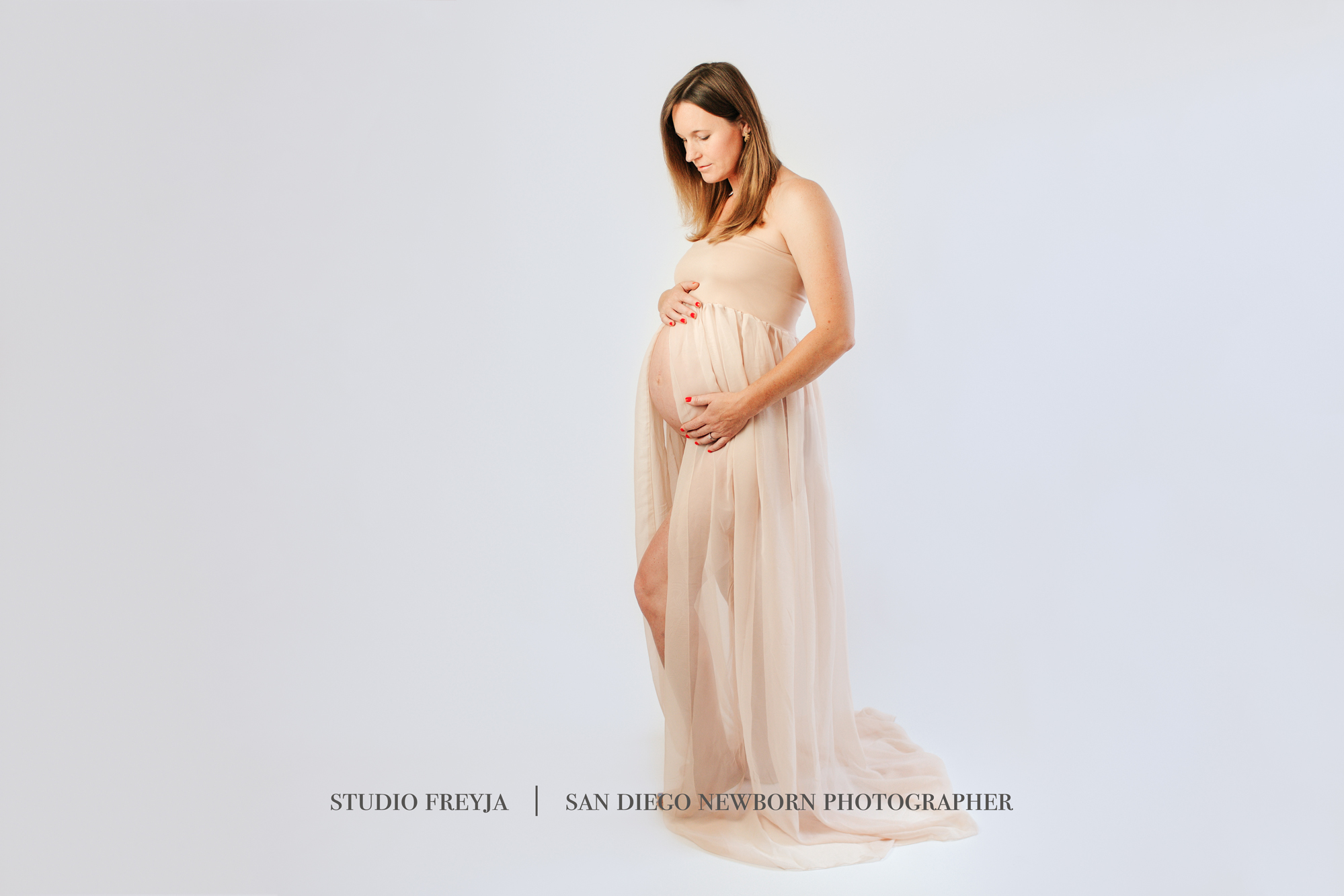 Pregnant woman in a sheer blush gown looking down at her bump on a light gray backdrop