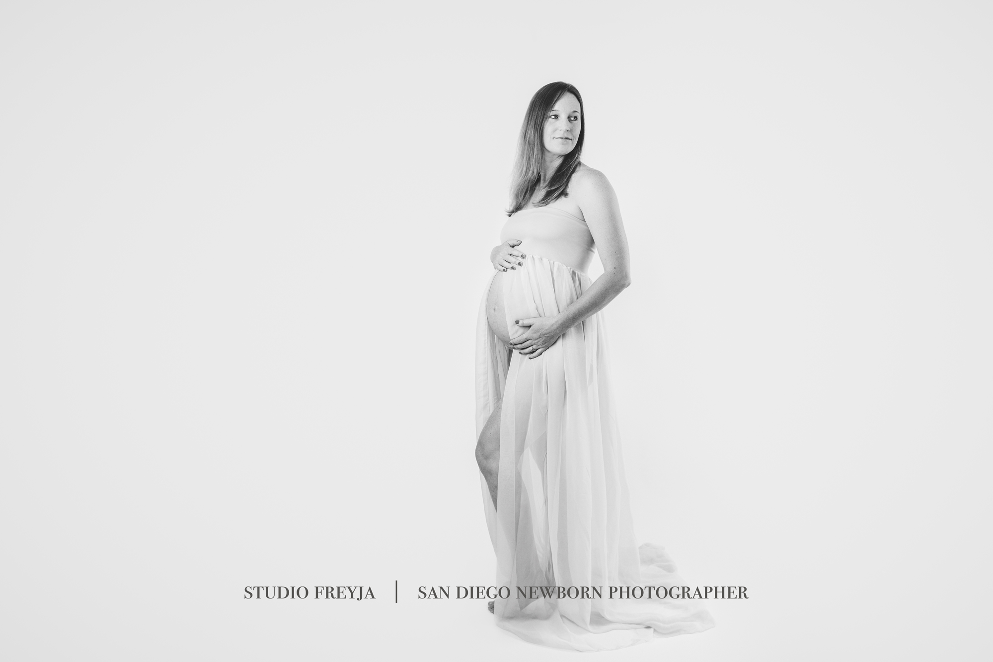 Black and white photo of a pregnant woman in a sheer gown standing in profile in a white studio