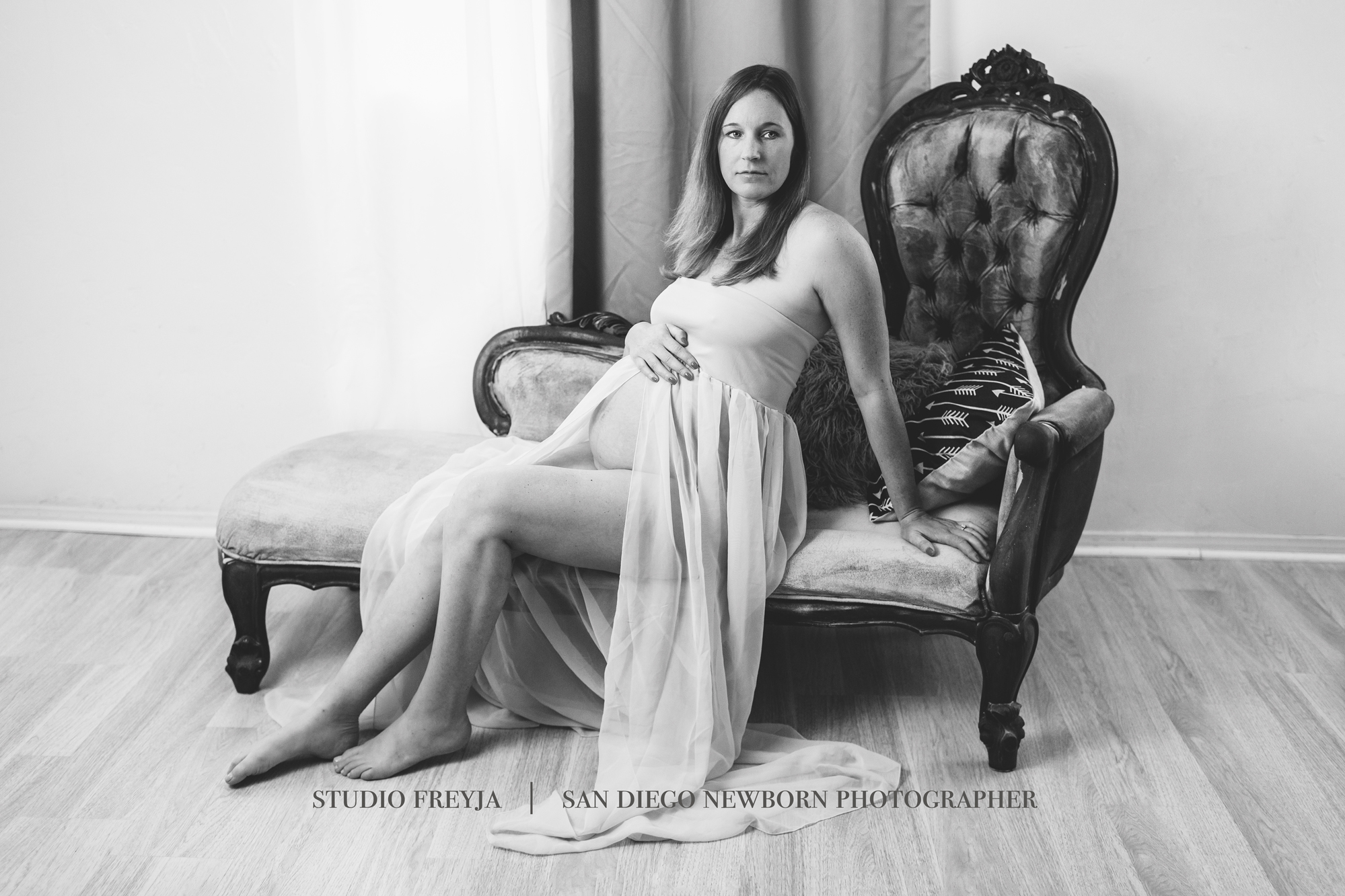 Black and white photo of a pregnant woman seated on an antique chaise in a sheer gown