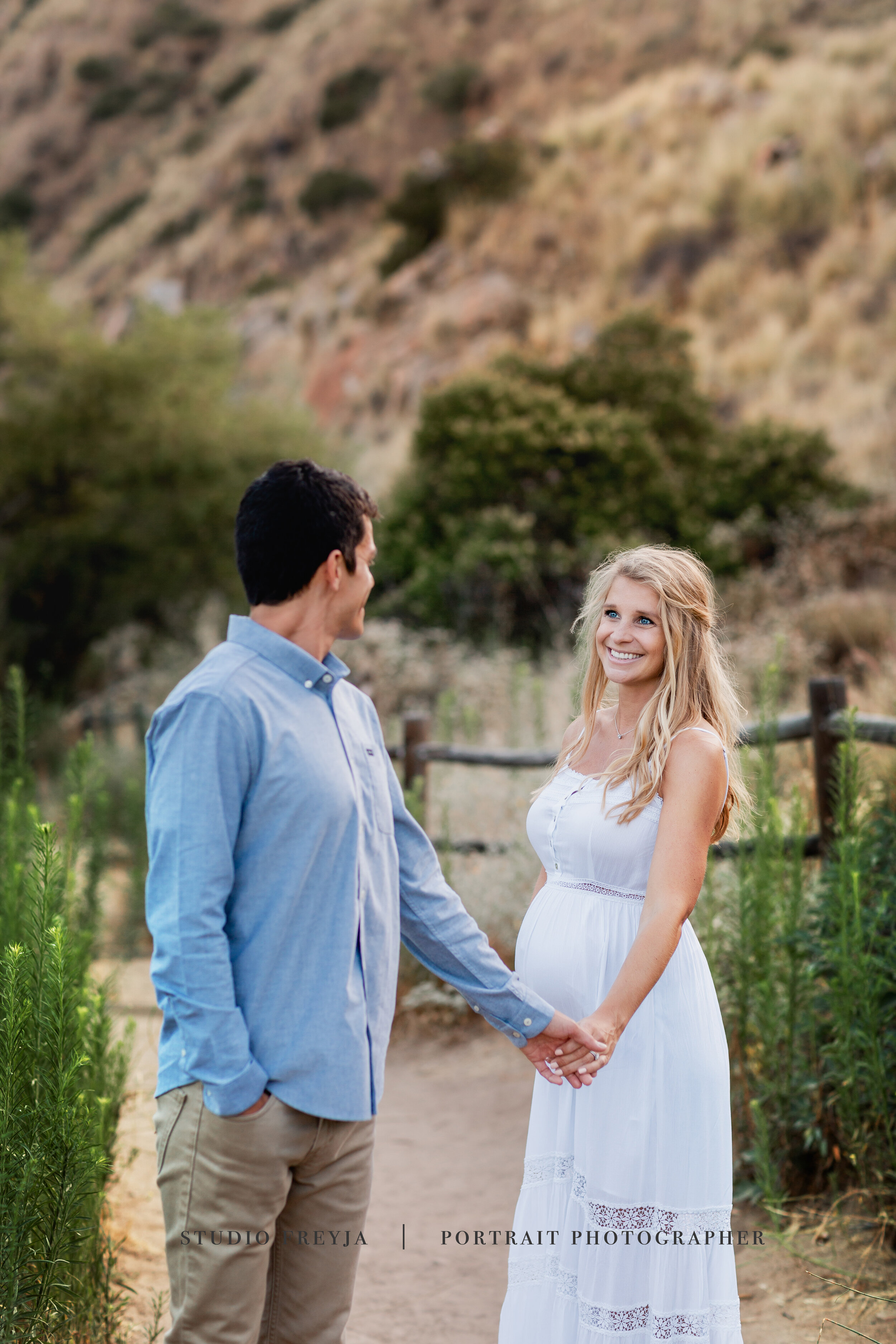 Expectant couple holding hands and smiling at each other on a dirt trail
