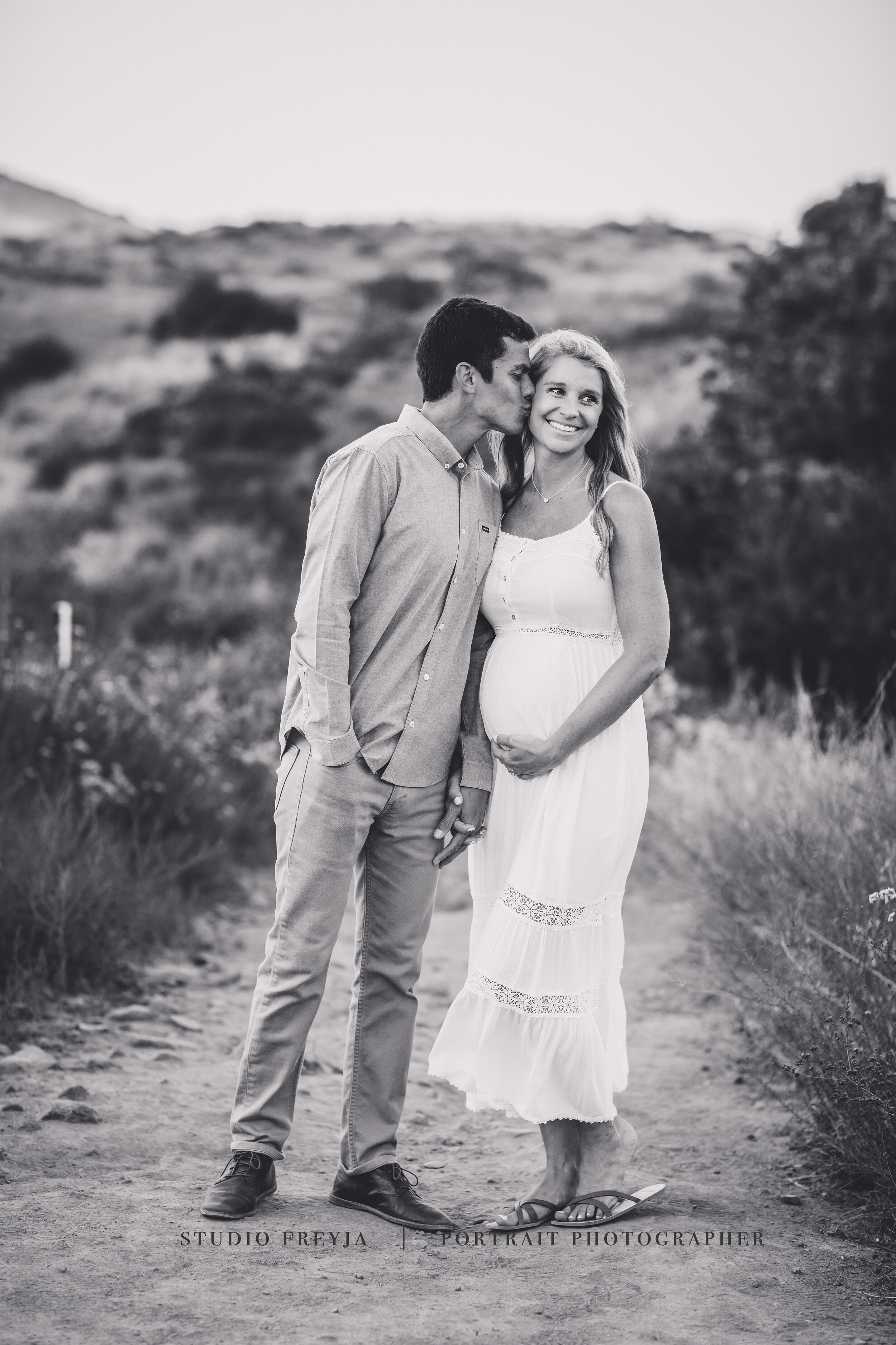Black and white photo of a husband kissing his pregnant wife's cheek on a trail