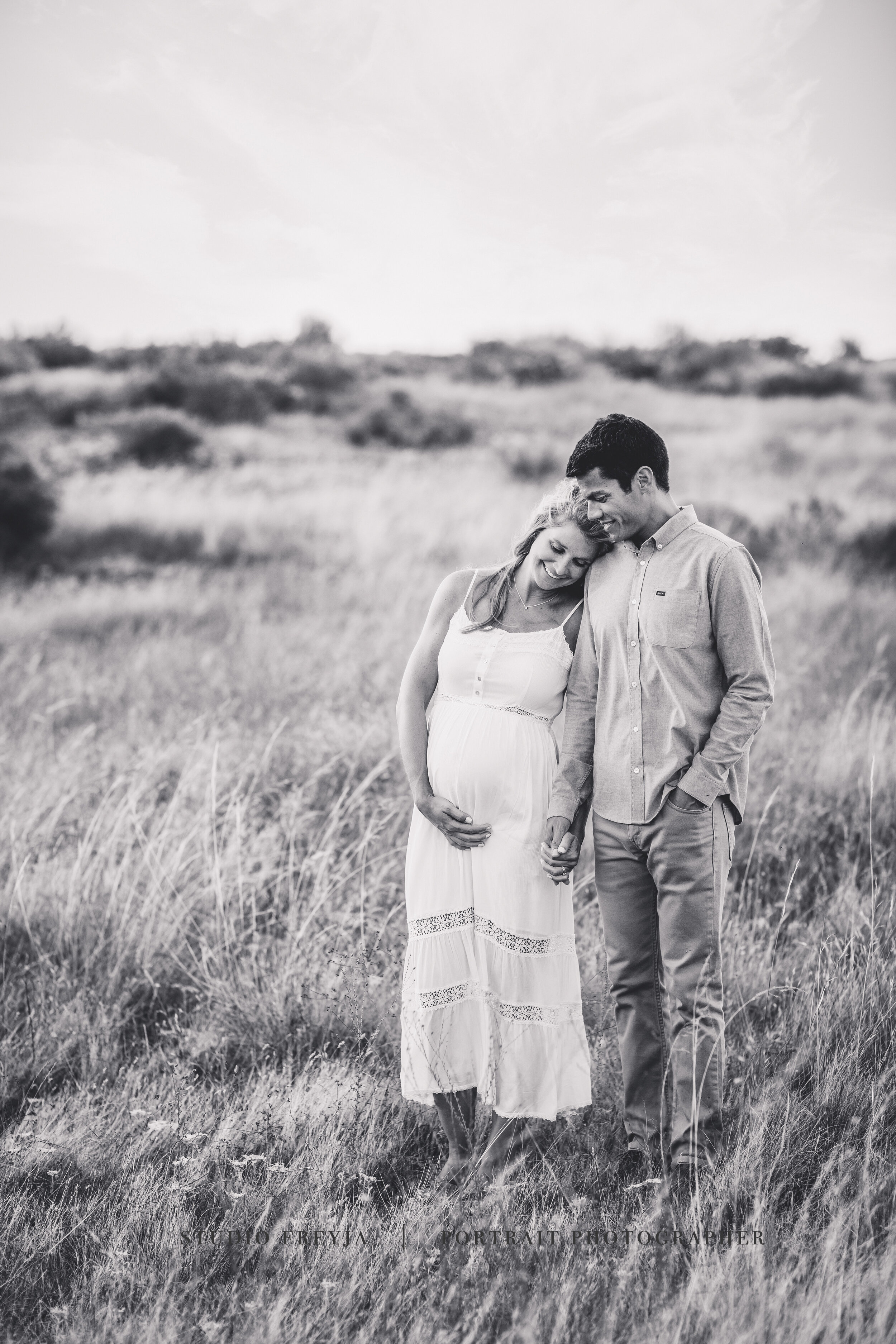 Black and white photo of a pregnant woman leaning her head on her husband's shoulder in a meadow