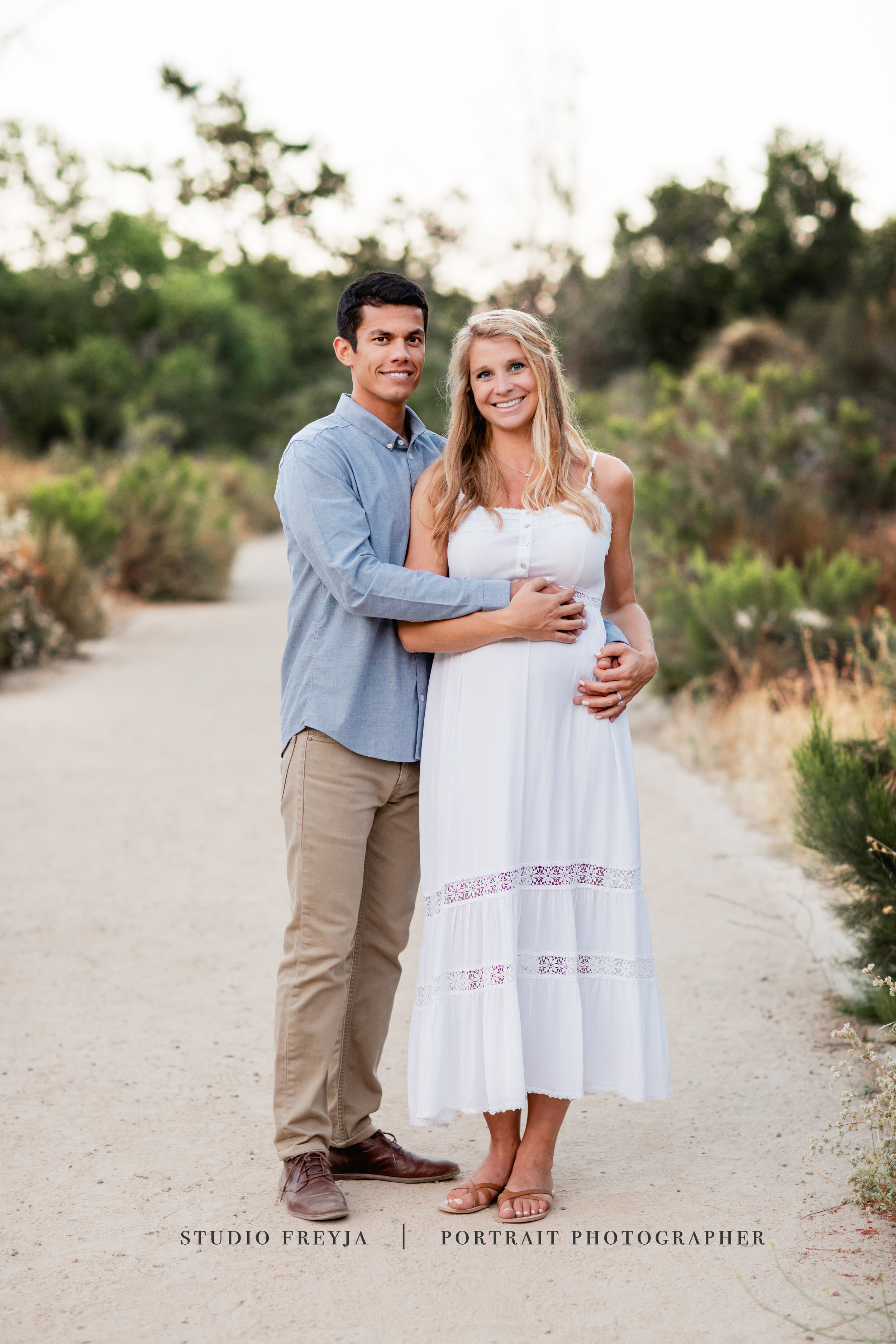 Husband holding his pregnant wife from behind on a garden path, both smiling
