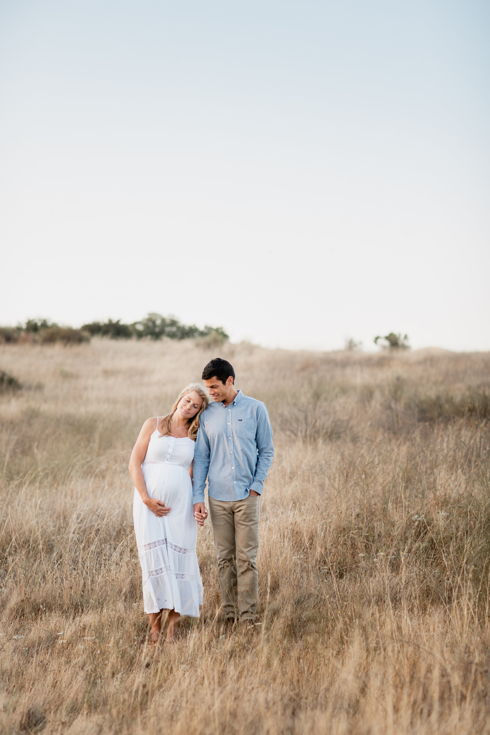 Expectant couple leaning into each other in an open field at golden hour