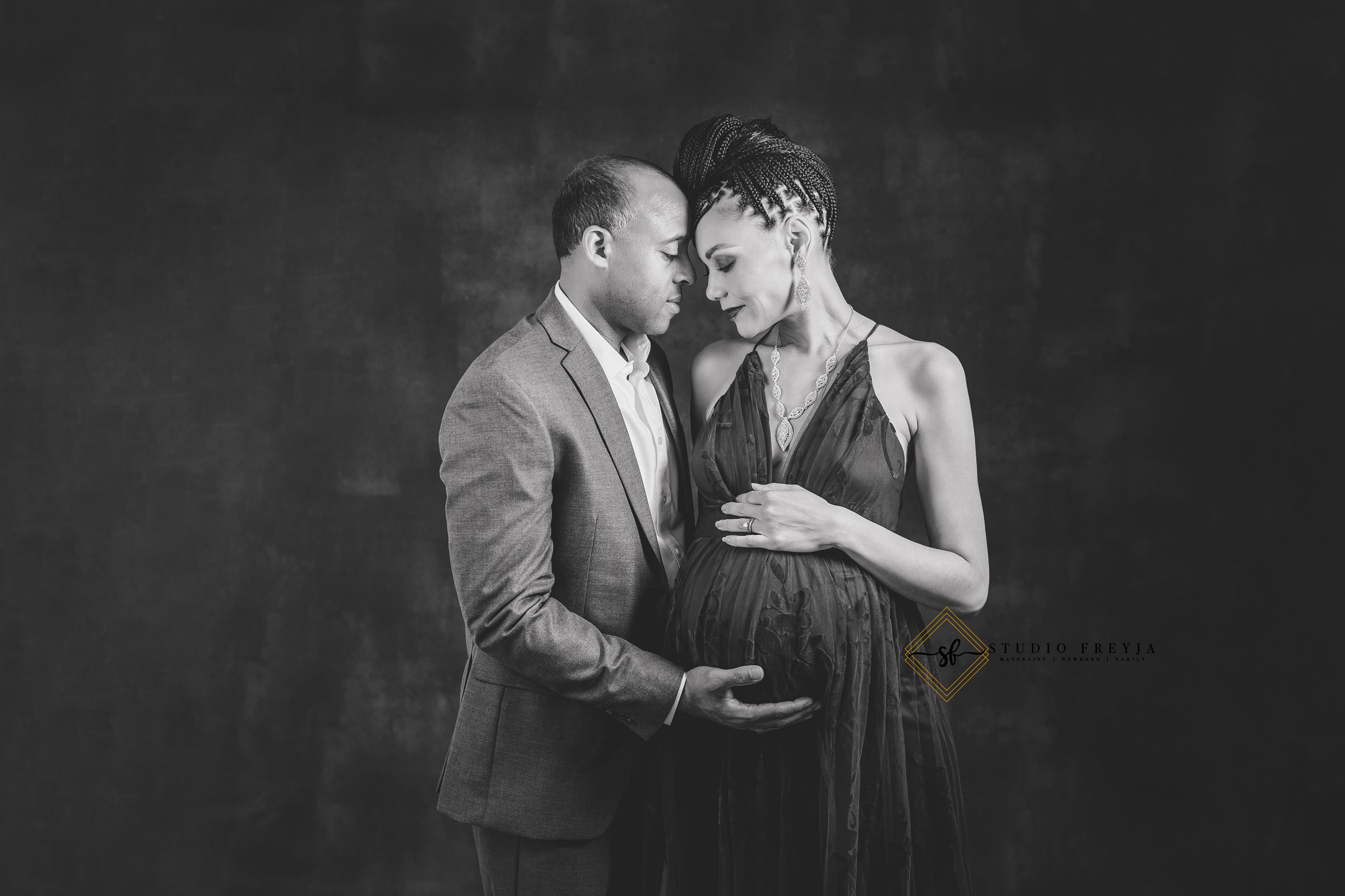 Black and white studio photo of an expectant couple forehead to forehead, his hand on her bump