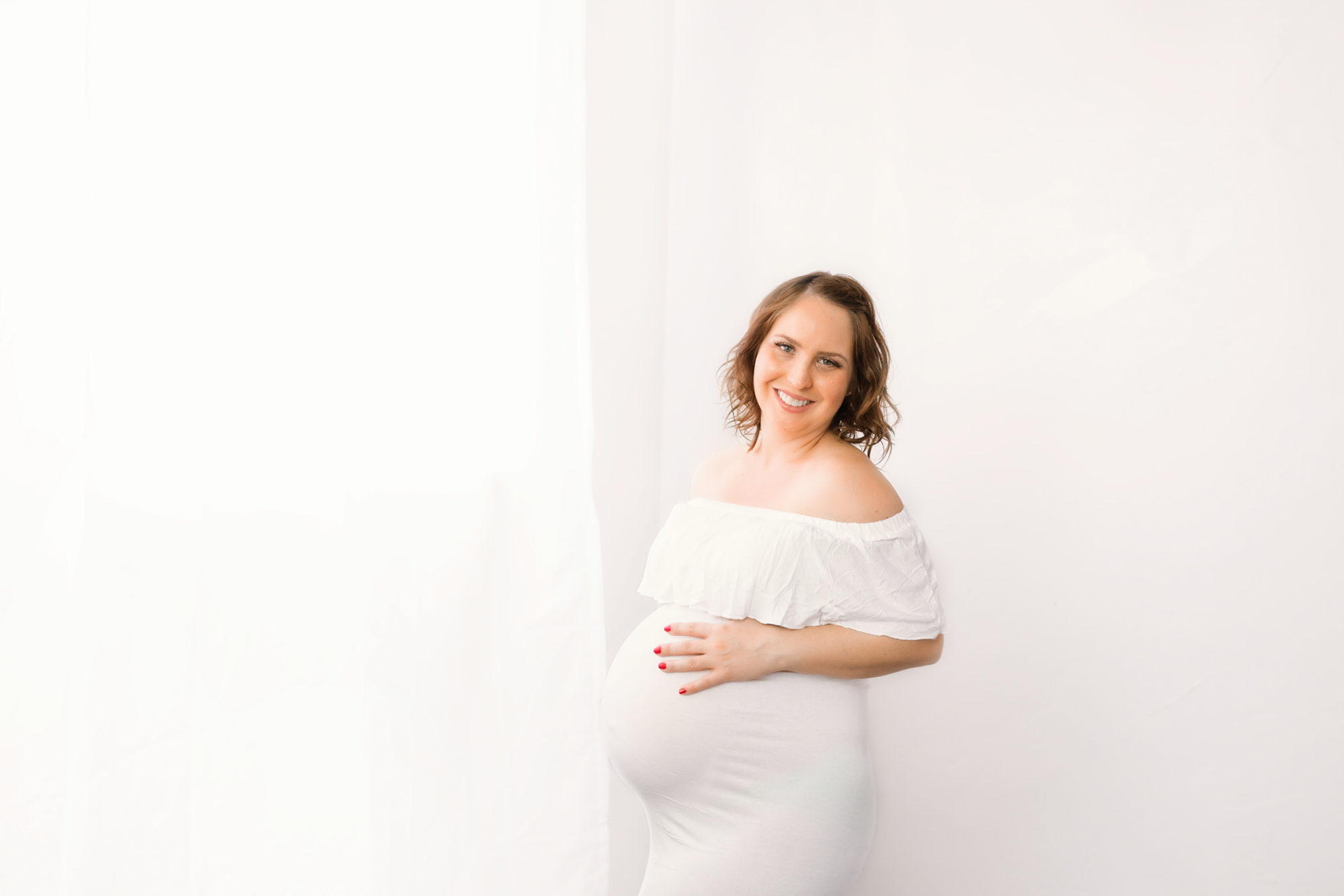 Smiling pregnant woman in a white ruffled dress by a bright window