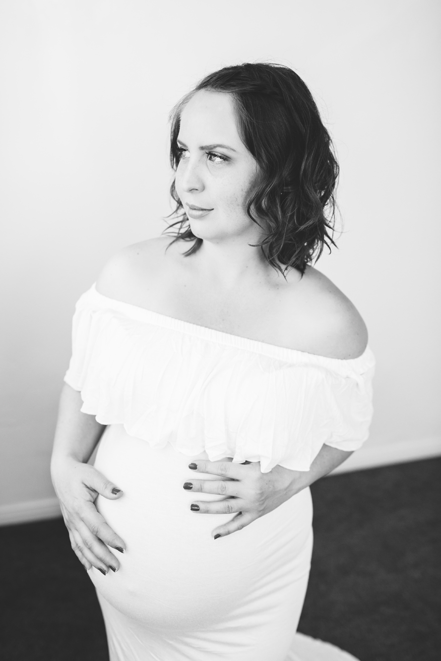 Black and white photo of a pregnant woman in a white off-the-shoulder dress looking over her shoulder