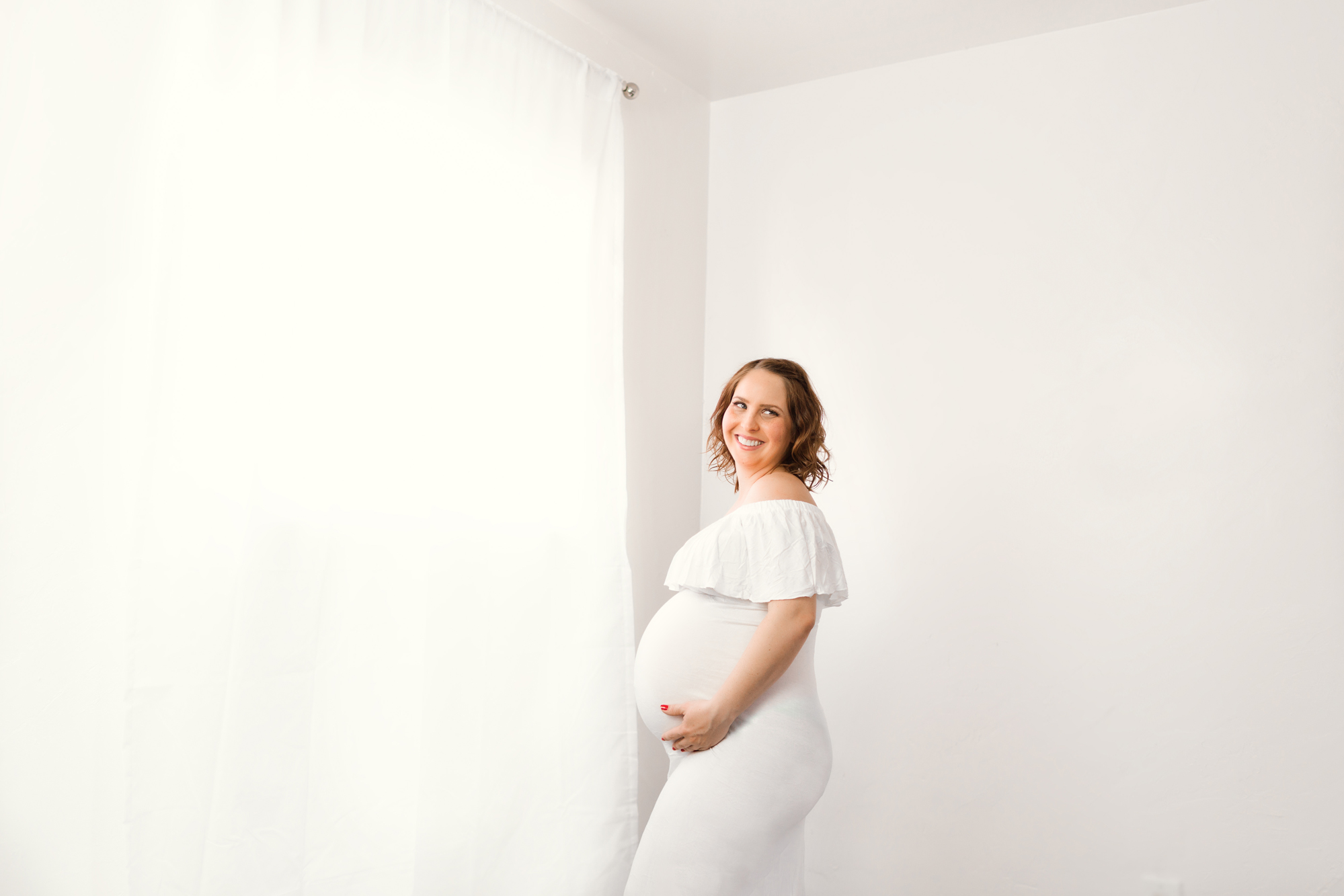 Smiling pregnant woman in a white ruffled dress standing by a sheer curtain in a bright studio