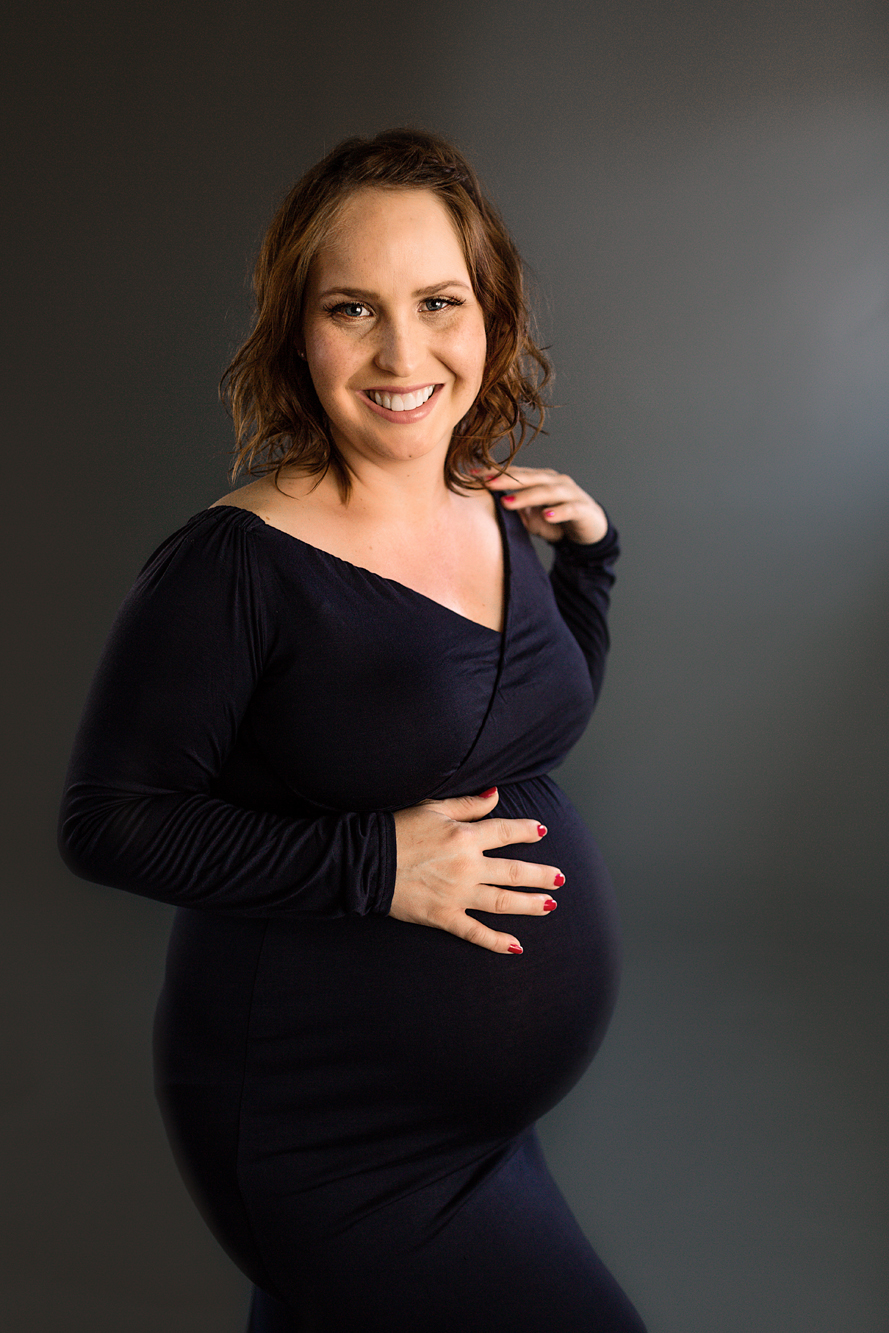 Smiling pregnant woman in a navy wrap dress cradling her bump against a dark gray backdrop