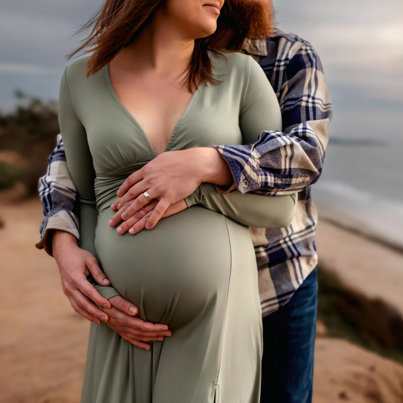 Close-up of a husband's arms wrapped around his pregnant wife's bump in a sage green dress on a coastal bluff